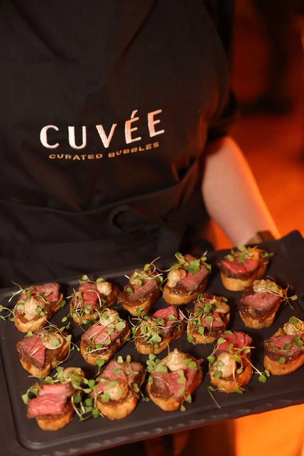 This close-up shot captures a server, identifiable by their black apron featuring the 'CUVÉE CURATED BUBBLES' logo, presenting a sleek matte black tray. The tray is laden with twelve meticulously arranged gourmet canapés, each a crostini topped with perfectly cooked rare beef, a creamy sauce, and vibrant microgreens. The background is softly blurred with warm, inviting orange ambient lighting, suggesting an upscale evening event or intimate dining setting.