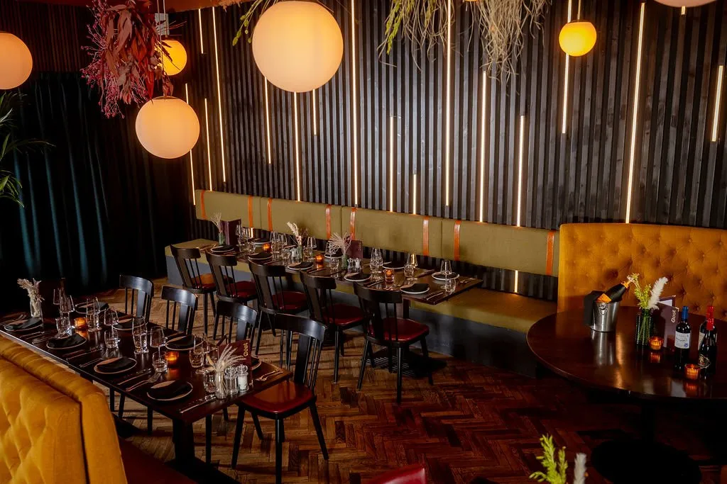 This intimate dining space features a sophisticated, moody ambiance, achieved through dim, warm lighting and rich material choices. Dark vertical slatted walls with integrated LED strips provide a striking backdrop to olive-green banquette seating accented with tan leather straps, complemented by plush mustard-yellow tufted booths. Dark wooden tables are meticulously set, while large spherical pendant lights and dried floral arrangements add a touch of organic elegance to the dark herringbone wood floor, creating an inviting and luxurious atmosphere.