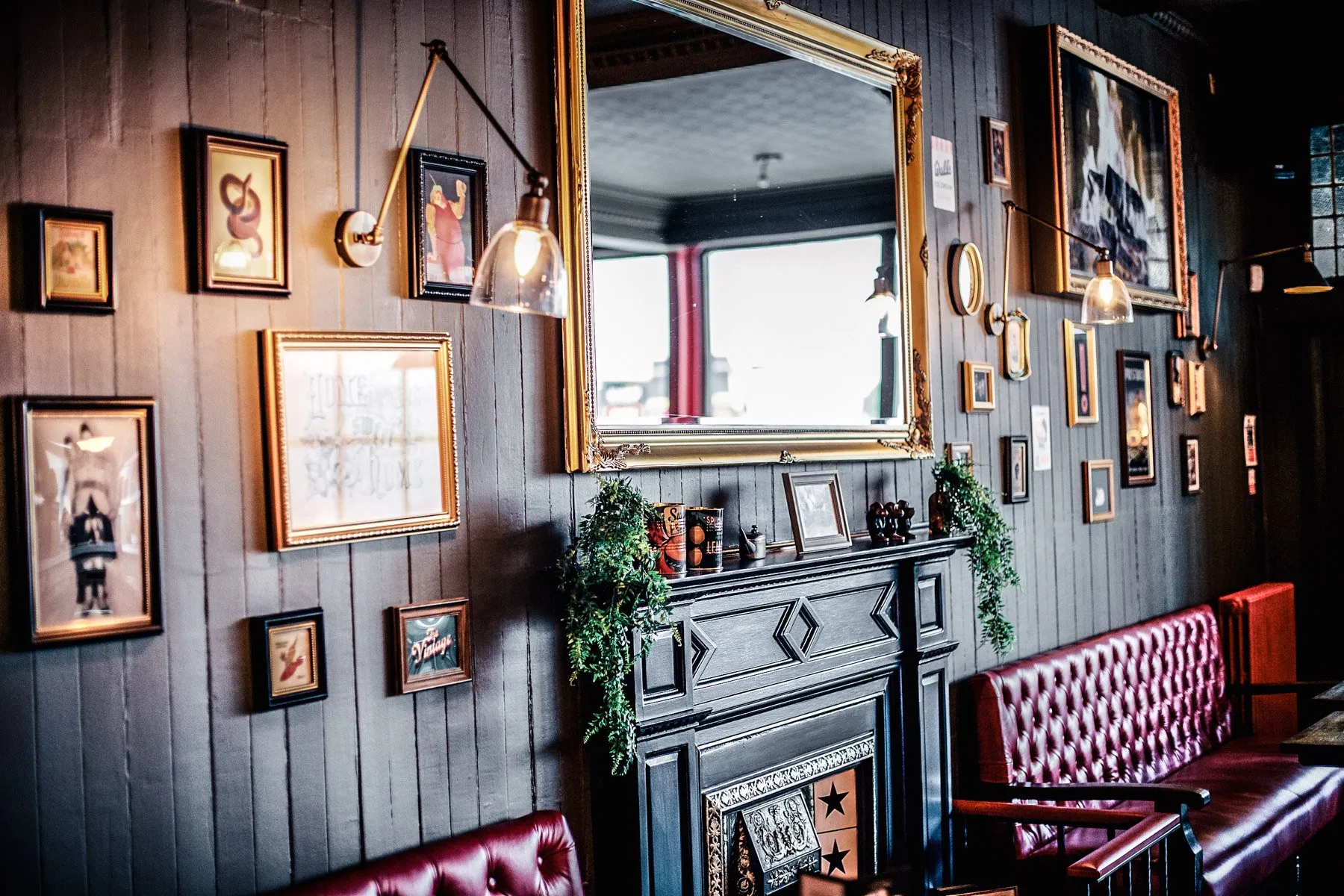 A richly textured and inviting interior features dark grey vertical wood paneling adorned with an eclectic gallery wall of framed art and a grand gold-framed mirror above a black, ornate fireplace. Deep red tufted leather banquette seating provides a luxurious and comfortable touch, illuminated by warm, directional sconces that cast a cozy glow, enhancing the traditional yet intimate atmosphere.