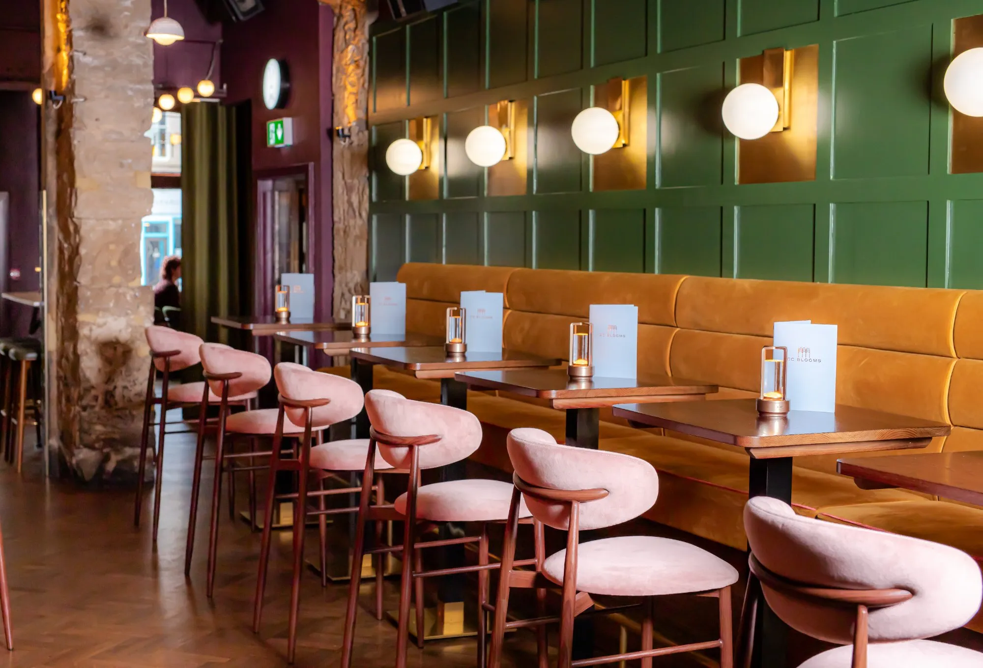 This inviting hospitality space features a rich material palette and warm lighting. A long, mustard-yellow velvet banquette runs along a dark green paneled wall, punctuated by elegant brass-backed spherical wall sconces. Opposite, dark wood tables are paired with plush, blush pink velvet bar stools, creating a sophisticated and intimate dining or lounge area. Exposed stone columns and a deep plum ceiling add texture and depth, while portable table lamps cast a soft glow on each setting.