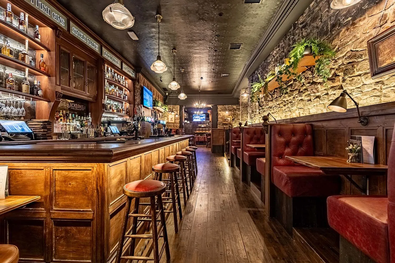 This inviting, long-format pub interior features a rich, traditional aesthetic. A substantial dark-stained wooden bar with matching stools runs along the left, backed by an ornate shelving unit displaying bottles under warm accent lighting. Opposite, a rugged exposed stone wall is softened by integrated wooden planters with greenery, complementing a series of deep red tufted leather booths and small wooden tables, all illuminated by a mix of classic pendant lights and task-style wall sconces, creating a cozy and intimate atmosphere.