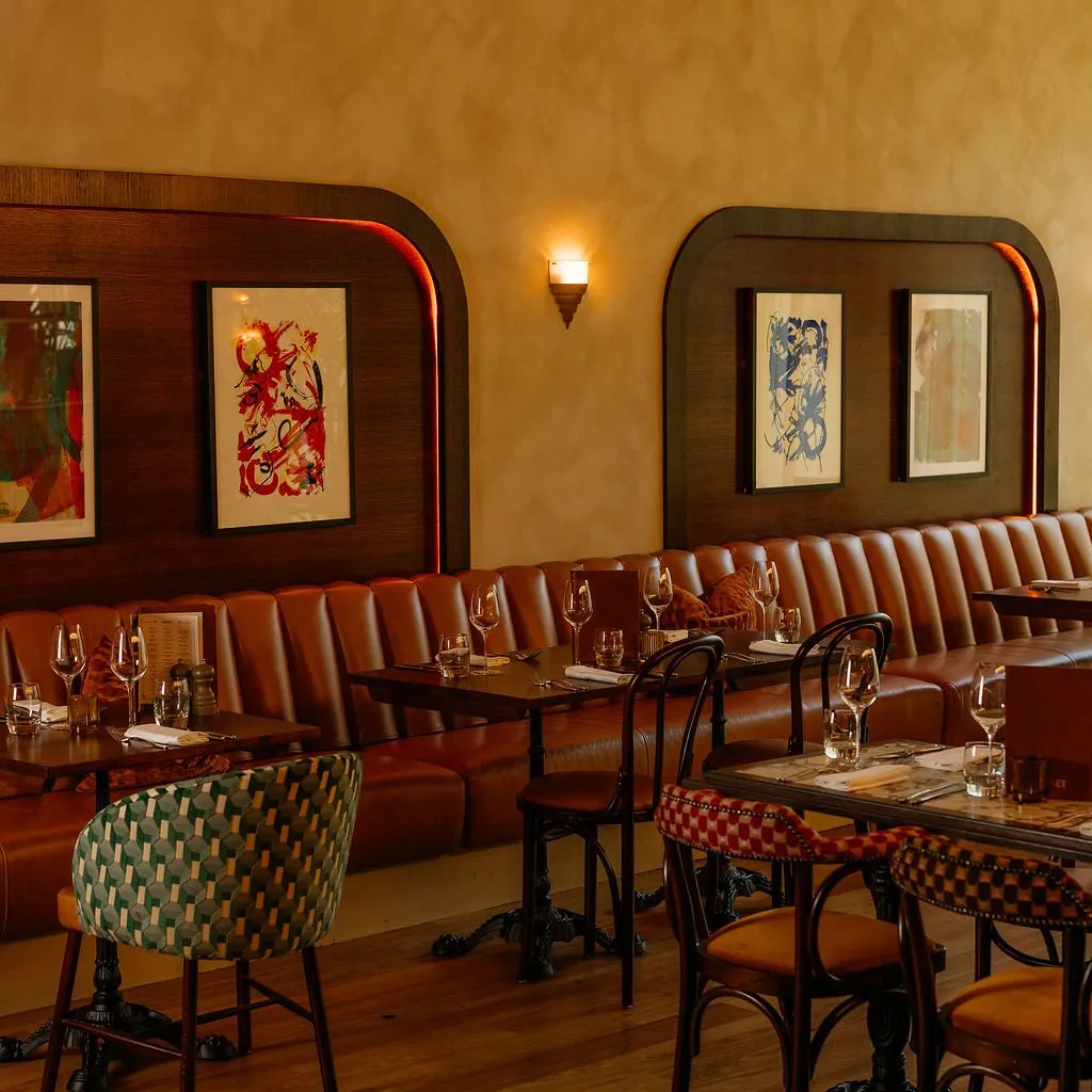 This inviting restaurant interior features a long, tufted tan leather banquette running along a textured ochre wall, framed by dark wood panels with integrated red LED strip lighting and abstract artwork. Square and rectangular dark wood tables with ornate cast iron bases are paired with an eclectic mix of dining chairs, including one with a green geometric pattern and others with a red checkerboard design. The warm, ambient lighting from a classic wall sconce and concealed LEDs creates a cozy and intimate atmosphere.