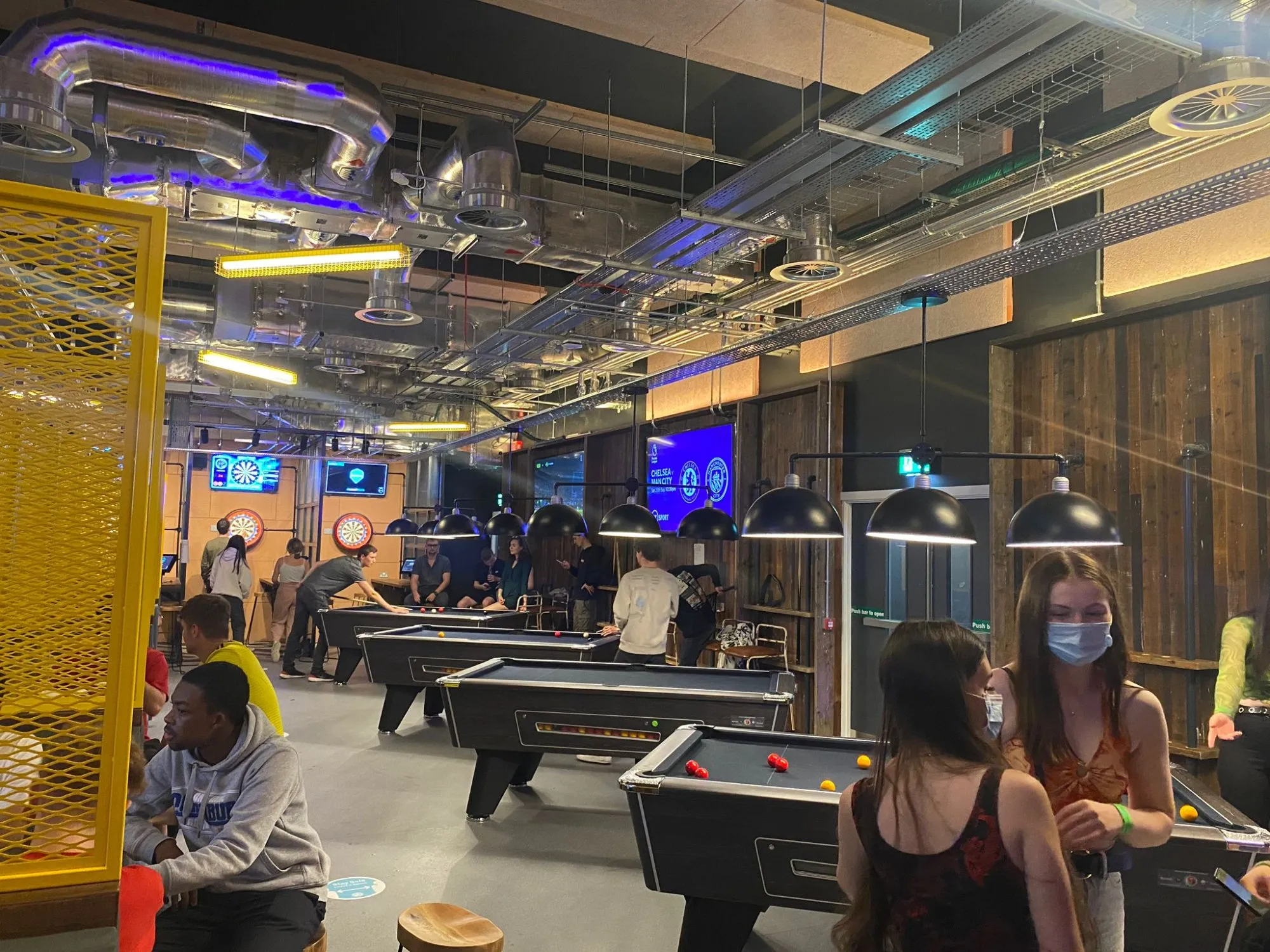This vibrant, industrial-chic entertainment space features multiple pool tables and dartboards, creating an energetic social hub. Exposed silver ductwork and cable trays dominate the ceiling, accented by cool blue LED lighting and linear yellow fluorescent fixtures, contrasting with warm raw timber wall cladding. The layout encourages interaction with various activity zones, while black dome pendants illuminate the pool tables, contributing to a dynamic yet intimate atmosphere.