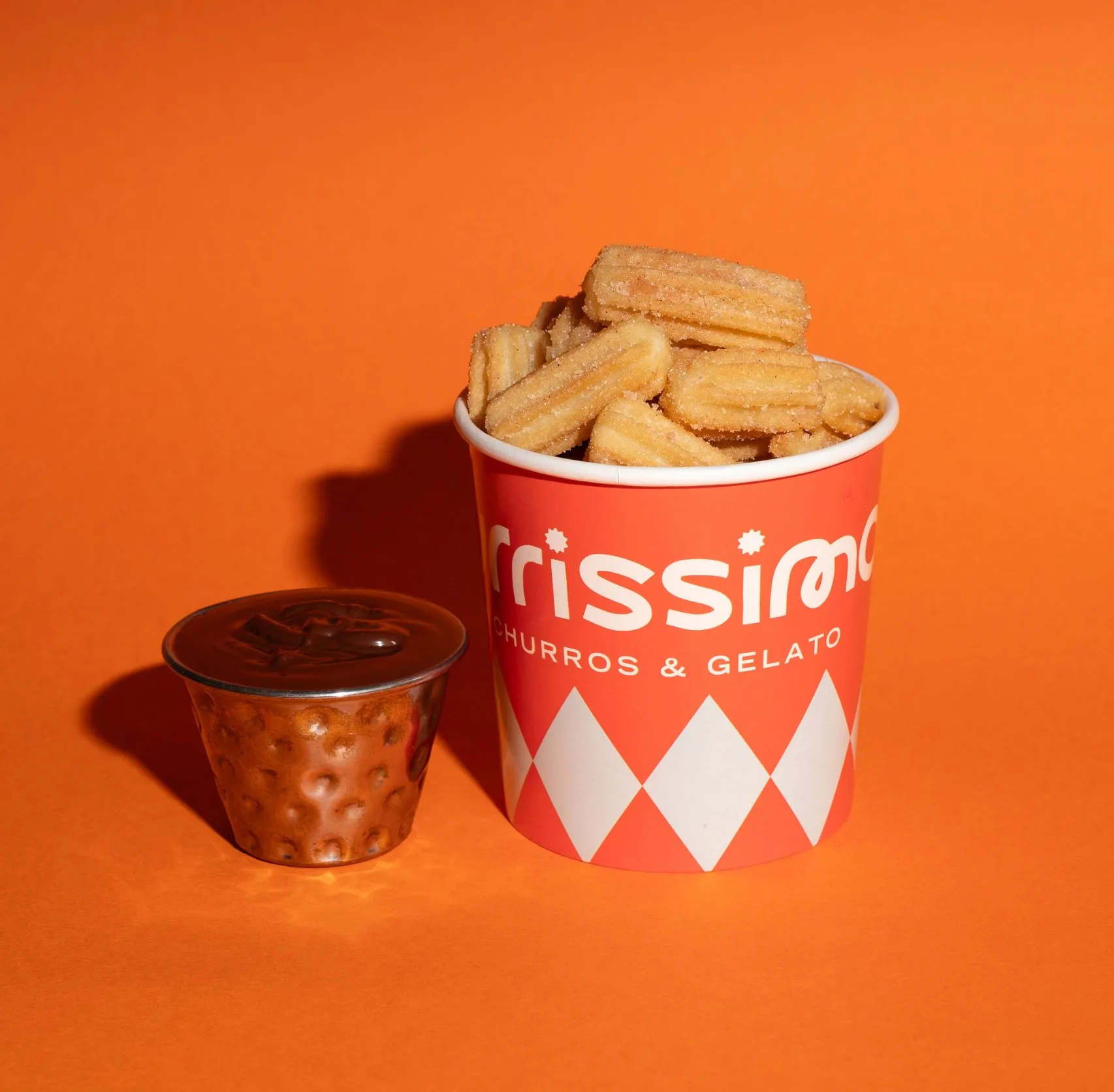 A vibrant, studio-lit close-up features a generous serving of golden, sugar-dusted churros in a branded red and white diamond-patterned paper cup, accompanied by a small hammered copper-toned metal cup filled with rich, dark chocolate dipping sauce. The items are set against a striking, solid orange background, creating a playful and appetizing scene with crisp shadows. This setup highlights the product's appeal and brand identity.