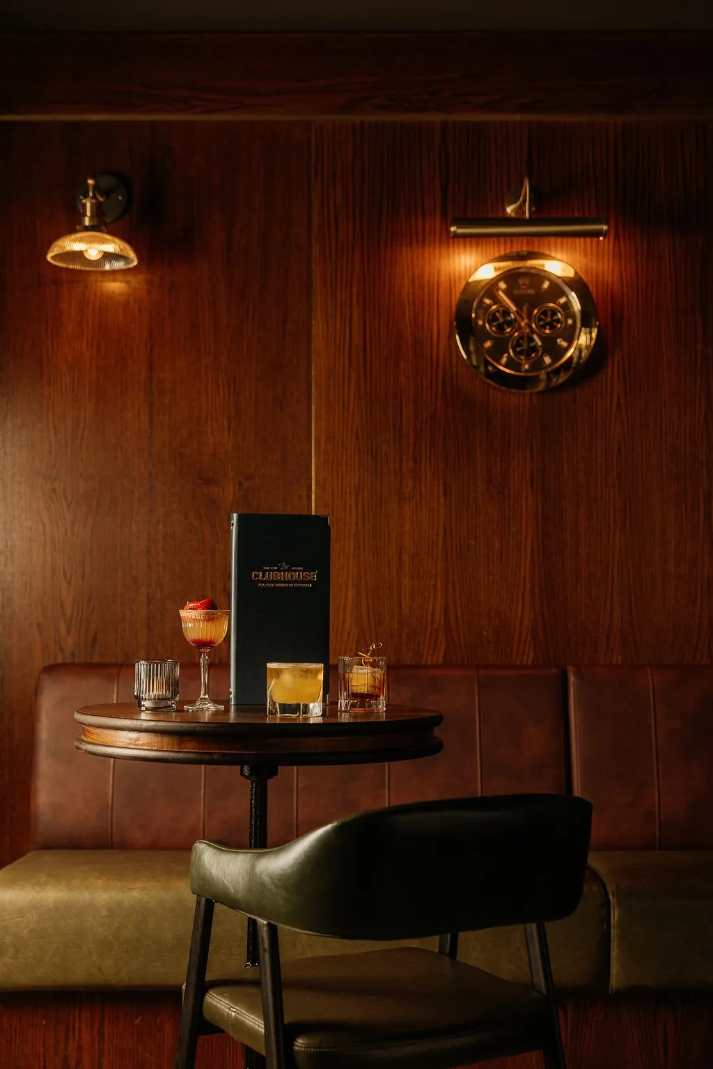 This intimate shot captures a sophisticated lounge or bar setting, featuring rich dark wood paneling that sets a warm, classic tone. A plush two-tone leather banquette in deep brown and olive green, paired with a matching dark green leather chair, invites guests to a small round wooden table laden with expertly crafted cocktails and a menu. The ambient lighting from brass wall sconces, including one illuminating a striking brass chronograph clock, creates a moody and inviting atmosphere perfect for evening drinks.