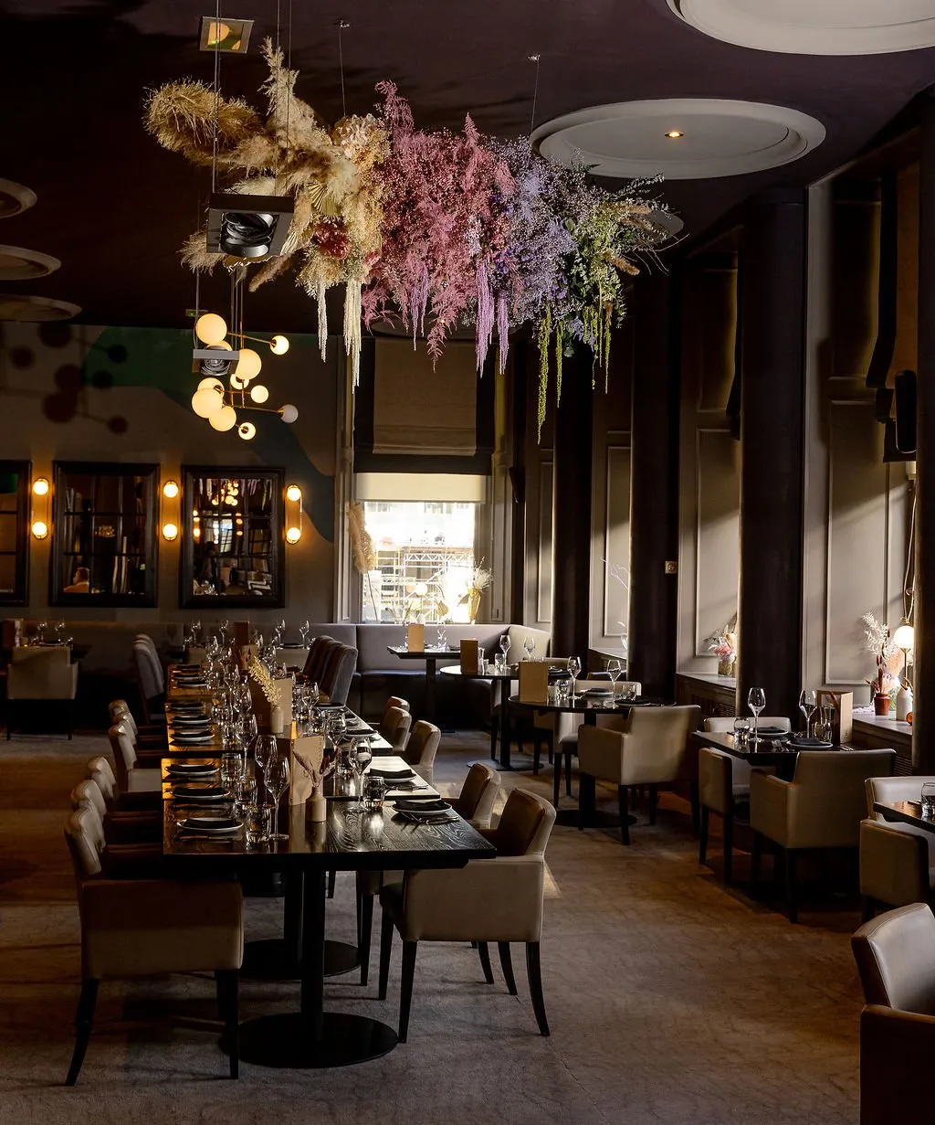A sophisticated dining room features a dramatic dried floral ceiling installation, transitioning from natural tones to vibrant pinks, purples, and greens, creating a striking focal point against the dark, moody interior. The space offers varied seating, including a long communal table with light upholstered armchairs and intimate banquette booths by tall windows, all set with dark wood tables. Warm, directional natural light streams through the windows, complementing the ambient glow from decorative wall sconces and a multi-globe pendant, enhancing the elegant and intimate atmosphere.