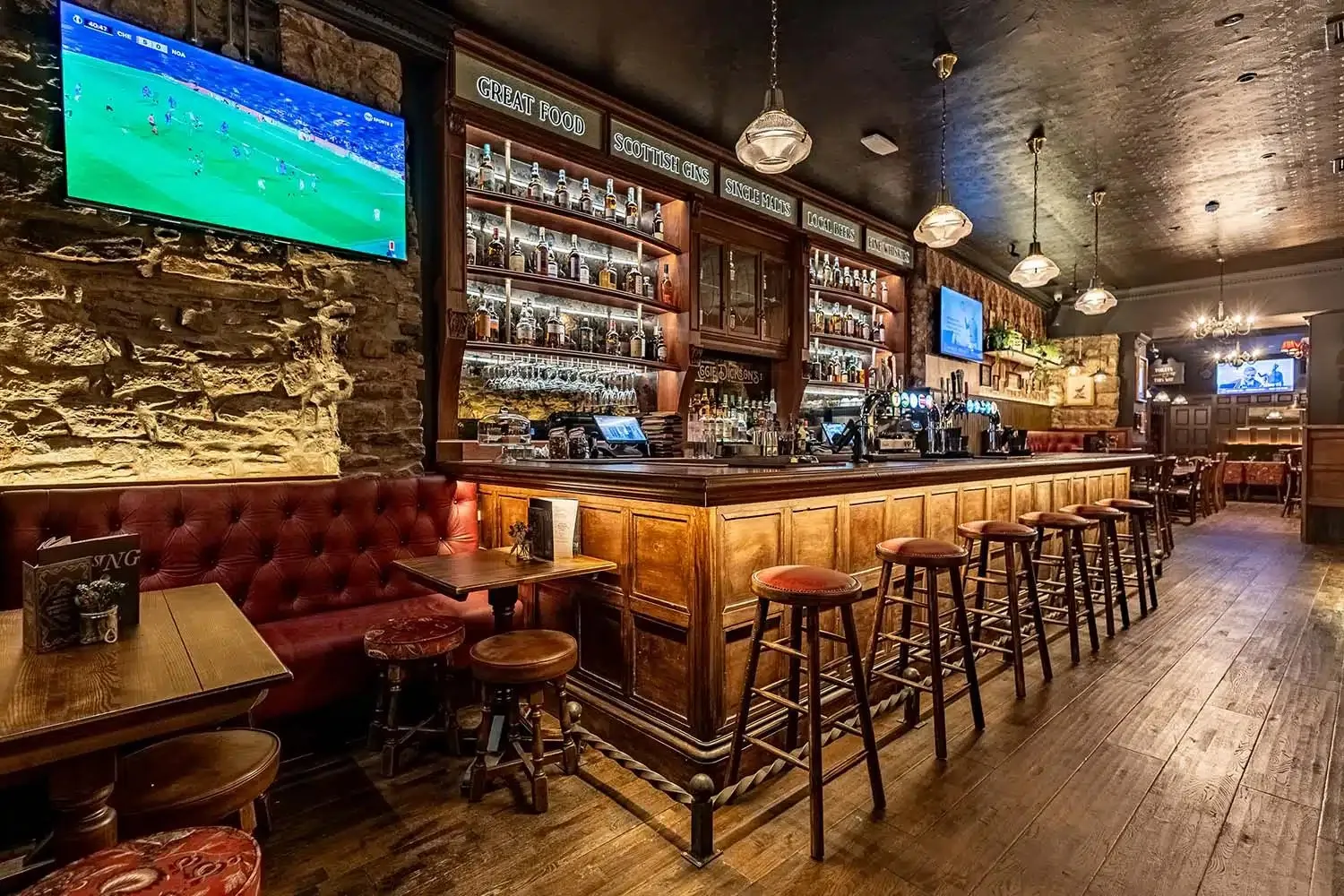This inviting pub interior features a long, richly detailed wooden bar counter with a comprehensive back bar display, illuminated to showcase an array of bottles. Opposite, a deep red tufted leather banquette runs along an exposed stone wall, creating cozy nooks with small wooden tables. The warm, dim lighting from traditional pendant fixtures and subtle backlighting cultivates a classic, convivial atmosphere, complemented by sturdy wooden bar stools and plank flooring, with additional dining visible in the background.