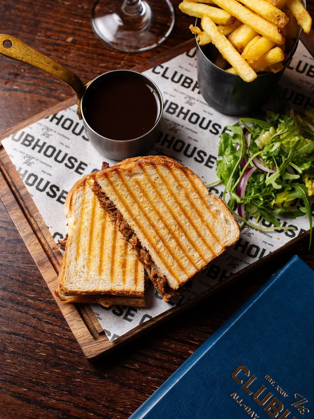 This close-up captures a well-composed table setting, showcasing a hearty meal that speaks to the venue's casual yet quality offering. A grilled sandwich, golden fries, and a fresh side salad are presented on a dark timber serving board, elevated by a branded paper liner, while a brass-handled pot of sauce adds a touch of rustic elegance. The rich, dark wood table and the deep blue menu with gold foil lettering reinforce a warm, inviting atmosphere, suggesting a sophisticated gastropub or casual dining space where attention to detail in presentation is key.