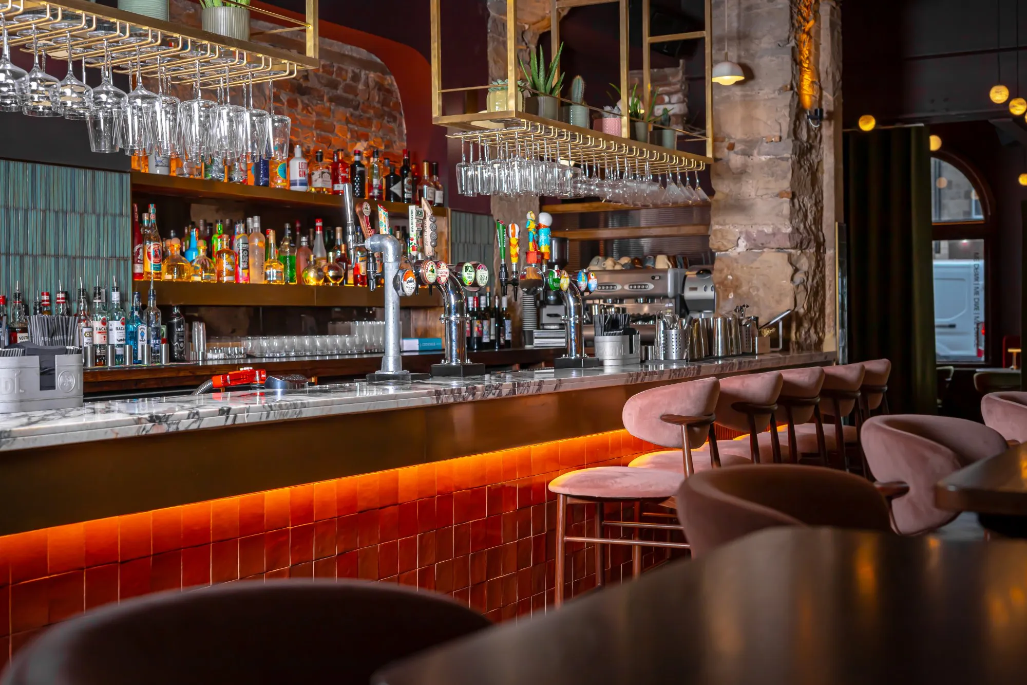This inviting bar scene features a sophisticated blend of textures and warm lighting. A long marble-topped bar with a brass-toned front is dramatically underlit, highlighting its terracotta-tiled base. Behind it, exposed brick walls and a raw stone column frame wooden shelves laden with bottles, while plush dusty-rose velvet bar stools offer comfortable seating, creating an intimate and luxurious atmosphere.