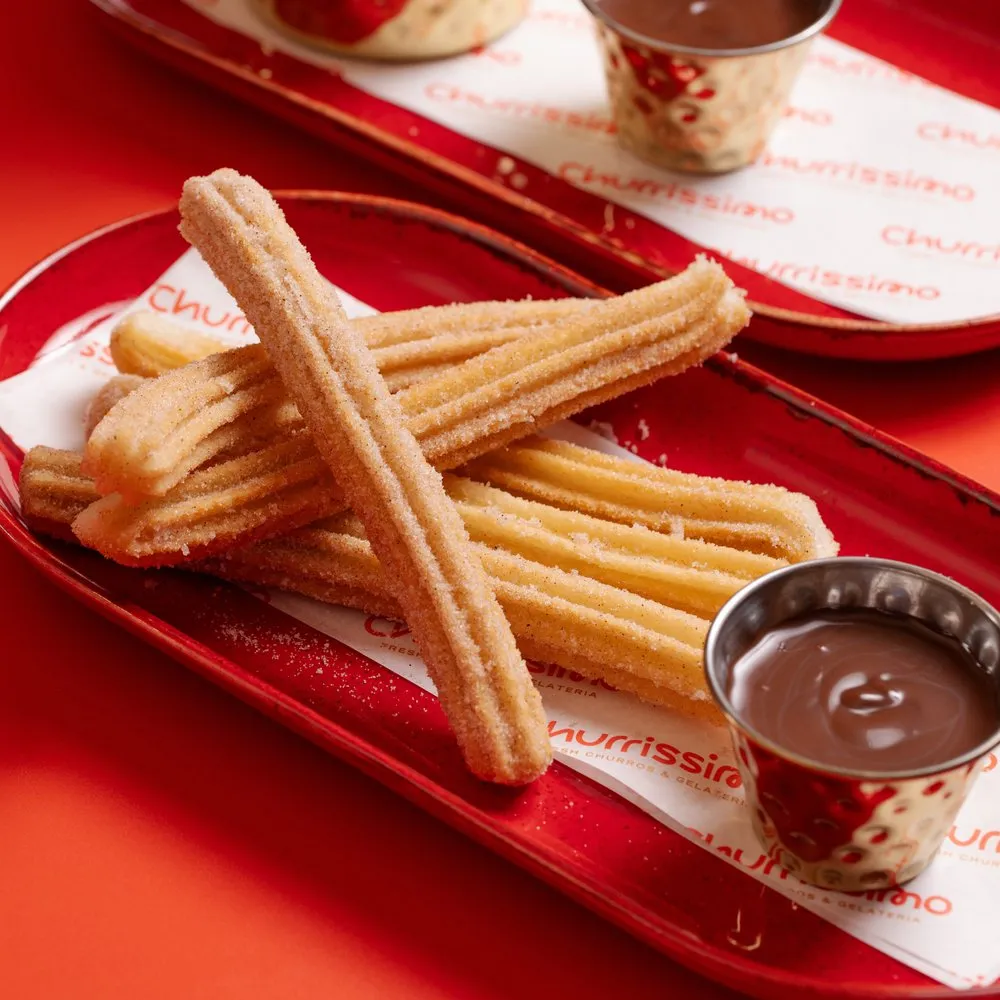 This close-up shot features a vibrant presentation of golden-brown churros, generously coated in cinnamon sugar, artfully arranged on a glossy, rectangular red ceramic plate. A small, textured metallic cup, possibly brass or copper, holds a rich, dark chocolate dipping sauce, adding a touch of rustic elegance. The branding "Churrissimo" is visible on the white paper lining the plate, all set against a warm, complementary red background that enhances the inviting and indulgent mood.