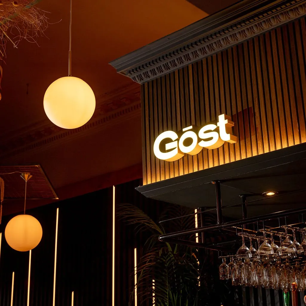 This close-up captures a sophisticated bar or restaurant interior, showcasing a blend of classic and contemporary design. A prominent backlit "Göst" sign is set against warm, vertical wood slat paneling, topped by an ornate, dark ceiling molding. The mood is intimate and inviting, illuminated by soft, spherical pendant lights and striking vertical LED strips, with a metal rack of wine glasses hinting at the bar's function.