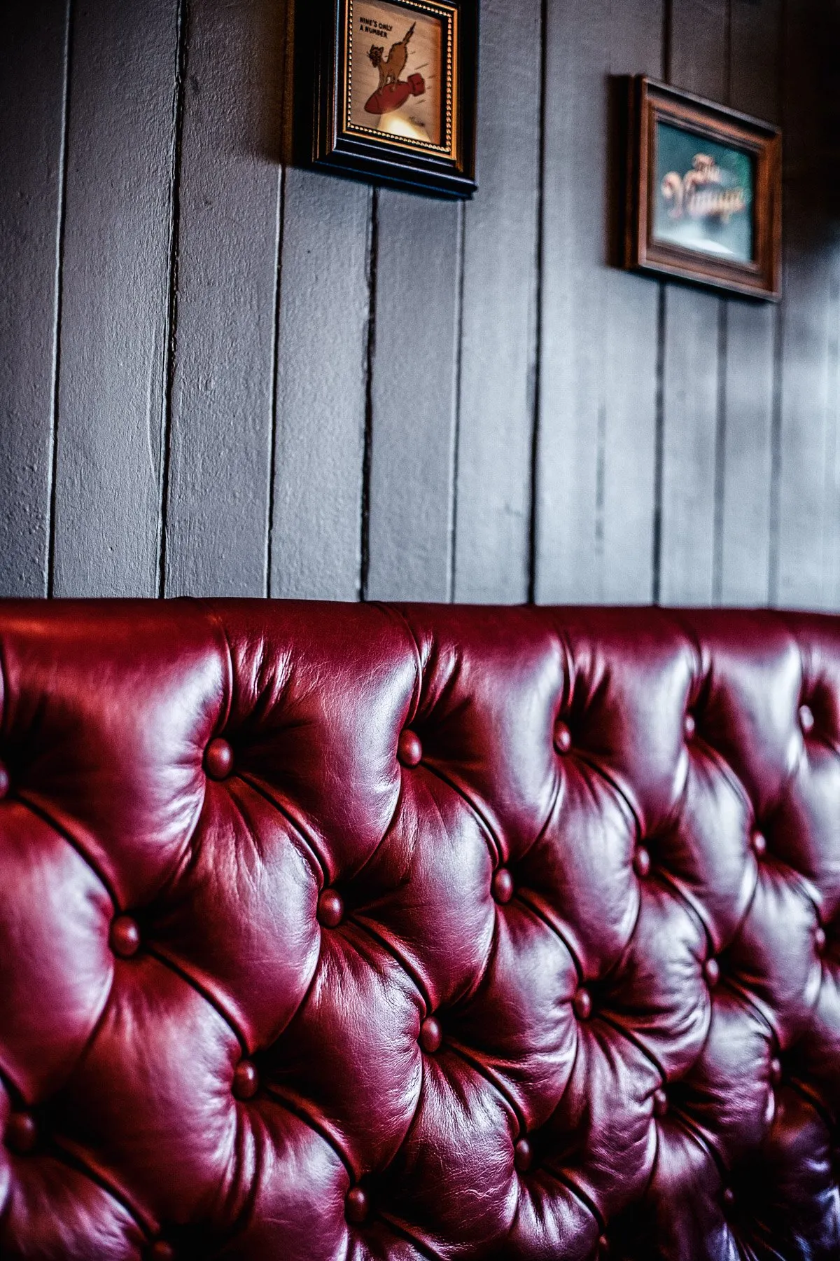 This close-up captures a luxurious, deeply tufted crimson leather banquette, its rich texture and sheen highlighted by soft, directional lighting. Behind it, a dark, vertically paneled wooden wall provides a sophisticated backdrop, adorned with small, framed vintage-style artwork that adds character and a hint of playful nostalgia to the intimate, moody setting. The overall impression is one of classic comfort and refined, old-world charm, perfect for a cozy lounge or upscale pub.