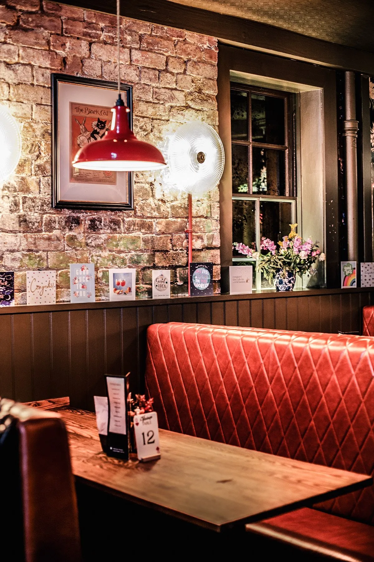 This inviting interior features a rich material palette, combining exposed brick walls with dark wood paneling and a striking red diamond-tufted banquette. Warm, ambient lighting from a vibrant red industrial pendant and wall sconces creates an intimate, classic pub atmosphere, complemented by a dark wooden table set for dining. Decorative elements like a vintage-style poster, displayed cards, and a vase of flowers add a personal, lived-in charm to the cozy corner, illuminated by the soft glow of evening through a multi-pane window.