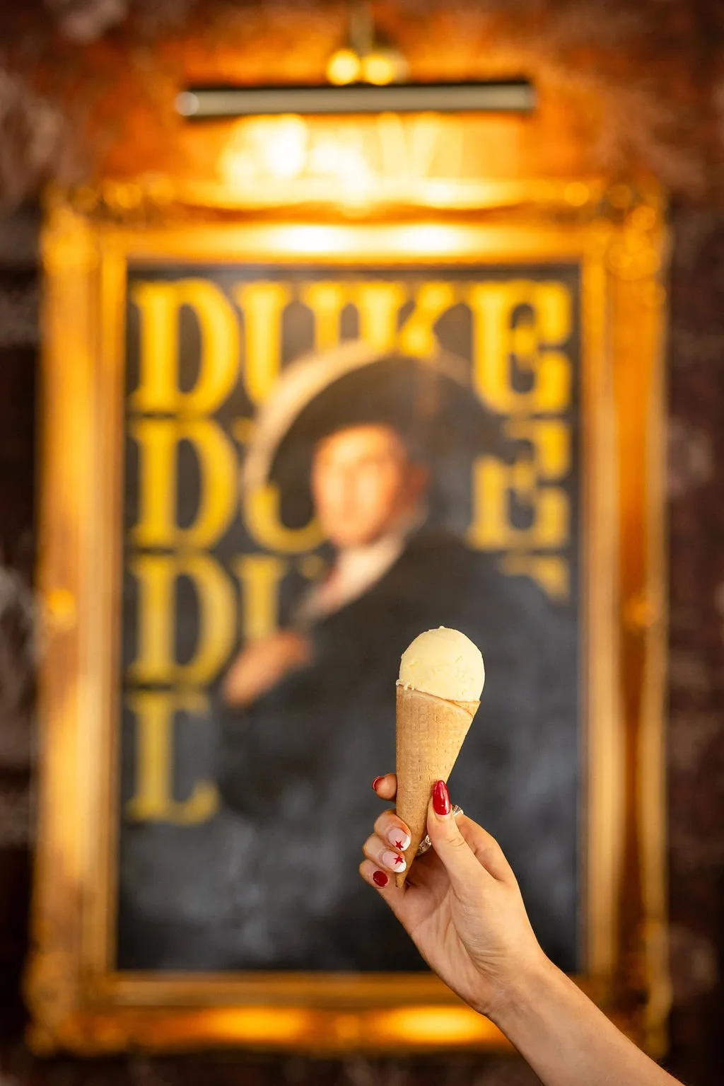 This close-up shot features a hand holding a classic vanilla ice cream cone, presented against a richly textured, dimly lit interior. In the blurred background, a large, ornate gilded frame highlights a contemporary artwork depicting a historical figure with bold yellow typography, creating a playful yet sophisticated contrast. Warm, ambient lighting emanates from above the frame, casting a golden glow that enhances the inviting and slightly whimsical atmosphere of the space.