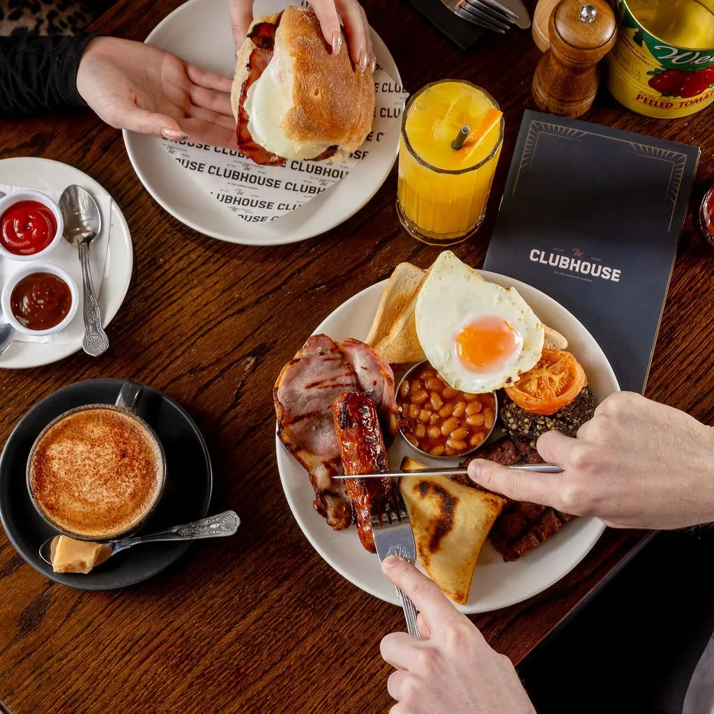 This overhead shot captures a cozy breakfast scene on a rich, dark wooden table, highlighting two patrons enjoying hearty meals. One plate features a full English breakfast with perfectly fried eggs, bacon, sausage, beans, and toast, while another holds a substantial bacon and egg roll. The warm lighting emphasizes the inviting atmosphere, complemented by a frothy cappuccino, fresh orange juice, and branded menu items. This suggests a casual yet quality dining experience, with attention to both the food and the overall guest comfort.