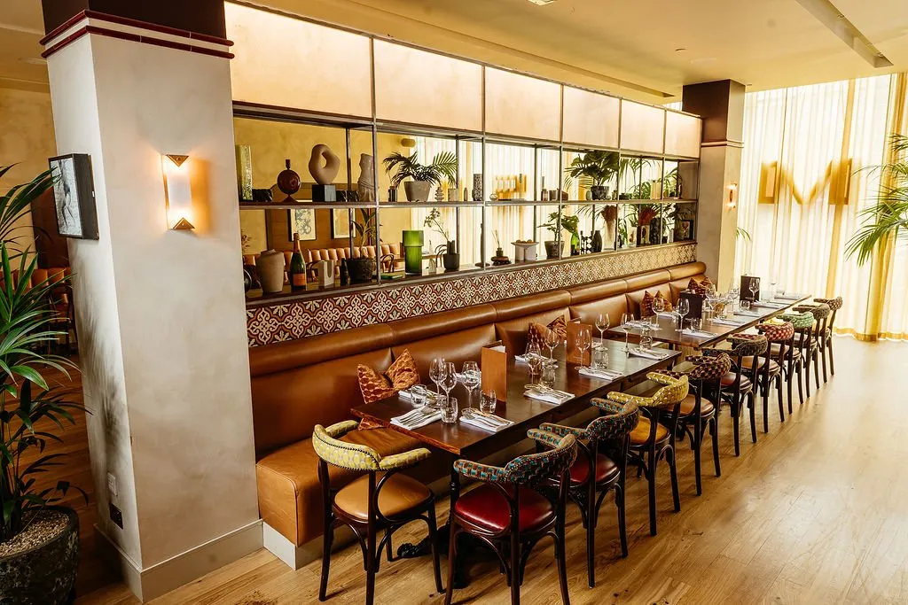 This inviting restaurant dining area features a long, continuous tan leather banquette running along one wall, paired with dark wood dining tables set for service. Above the banquette, a striking mirrored shelving unit displays an eclectic collection of plants and decorative objects, framed by a geometric patterned tile-like trim. The space is bathed in warm light from both natural sources filtering through sheer gold curtains and ambient wall sconces, creating a sophisticated yet comfortable atmosphere, further enhanced by the varied, patterned upholstery on the individual dining chairs.