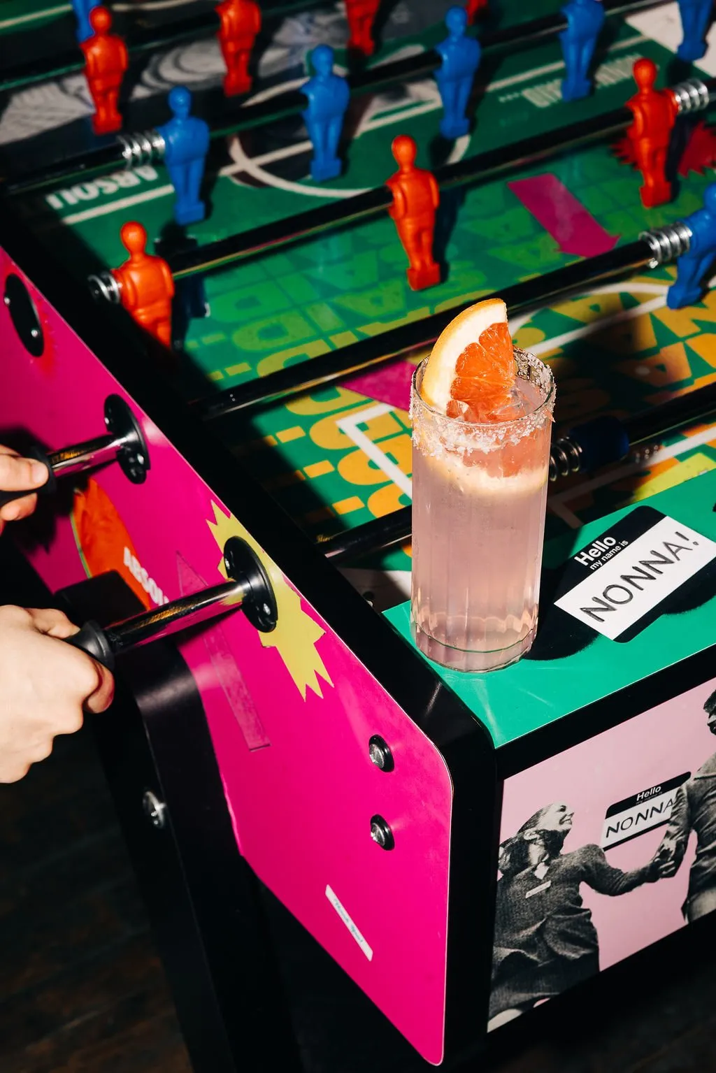 This close-up captures a lively foosball table, featuring bold graphic design in electric pink, vibrant green, and yellow, with classic red and blue players. A refreshing pink cocktail, garnished with a grapefruit slice and a salted rim, rests on the table's edge, while hands are actively engaged in play. The overall mood is energetic and playful, enhanced by the pop-art inspired graphics and the "Hello my name is NONNA!" sticker, suggesting a fun, casual hospitality setting.