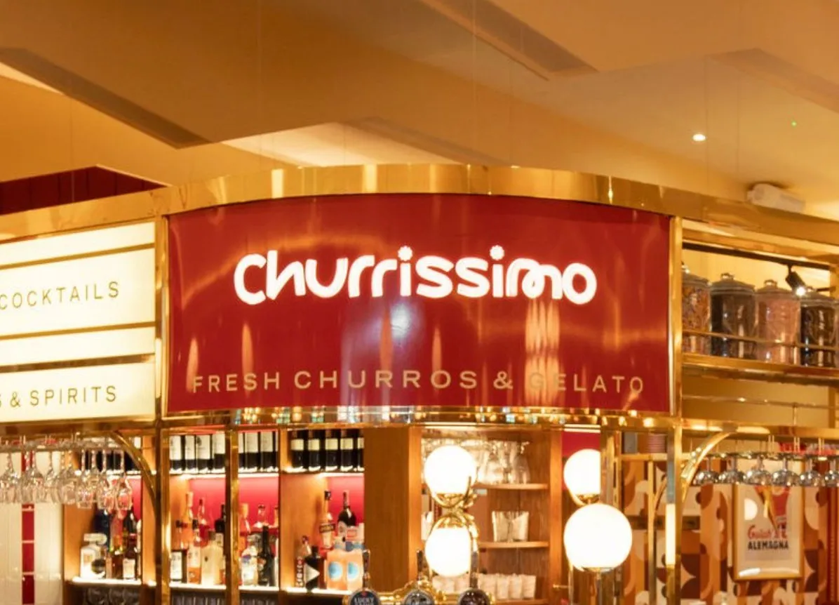 This close-up shot reveals a luxurious and inviting hospitality counter, featuring a prominent curved red sign for "Churrissimo" framed by polished brass. The space is richly detailed with warm, ambient lighting from elegant globe pendants and backlit shelving showcasing an array of spirits and decorative jars. The overall mood is sophisticated and slightly retro, emphasizing a premium experience for both churros/gelato and cocktails.