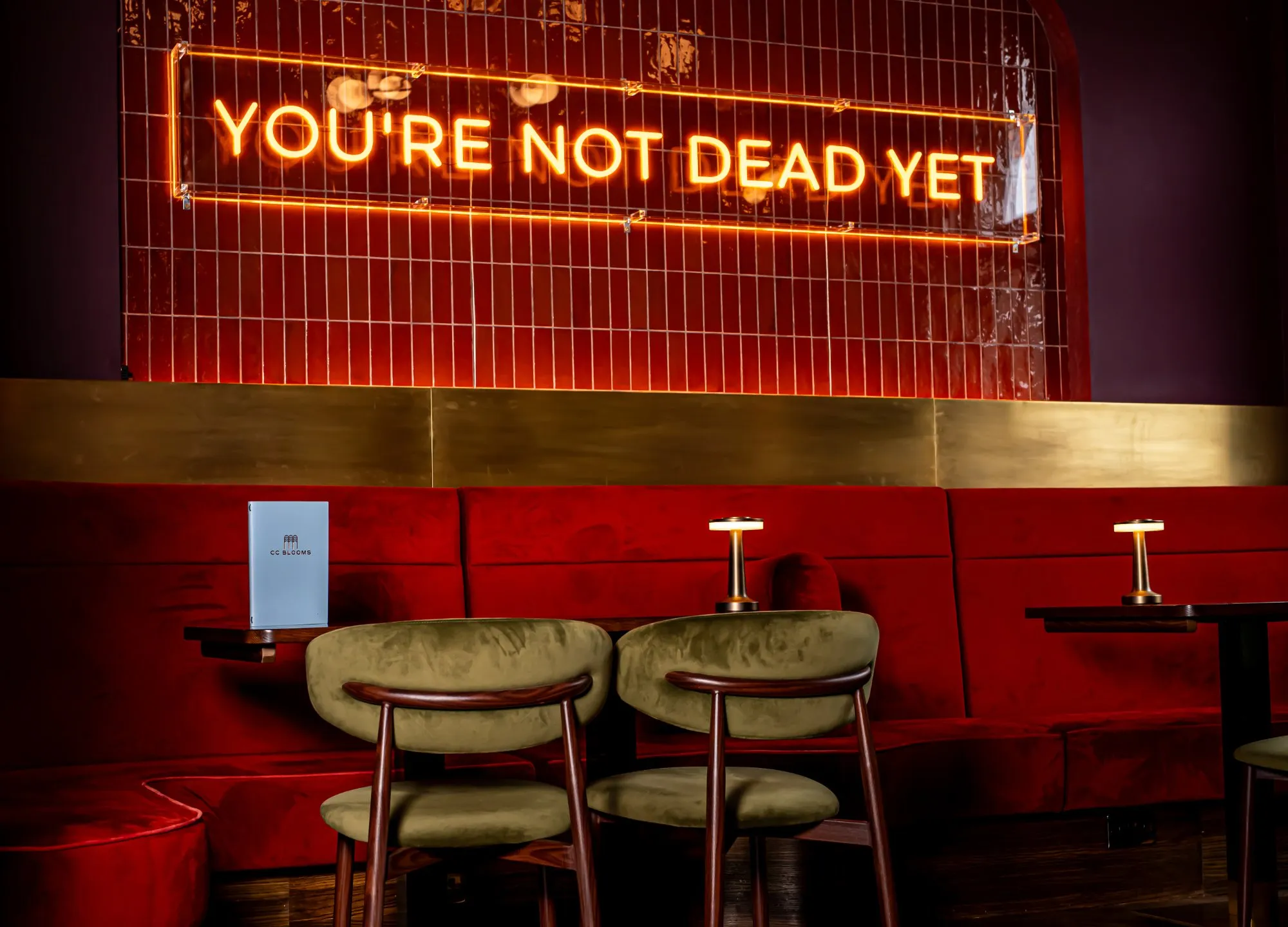 A vibrant, intimate seating area features a plush, deep red velvet banquette set against a wall of glossy, reddish-brown subway tiles. Above, a playful orange neon sign declares "YOU'RE NOT DEAD YET," casting a warm glow that highlights a brass-toned panel below. Olive green velvet chairs with dark wood frames complement the banquette, while small, modern table lamps on dark tables provide focused, ambient light, creating a sophisticated yet cheeky atmosphere.