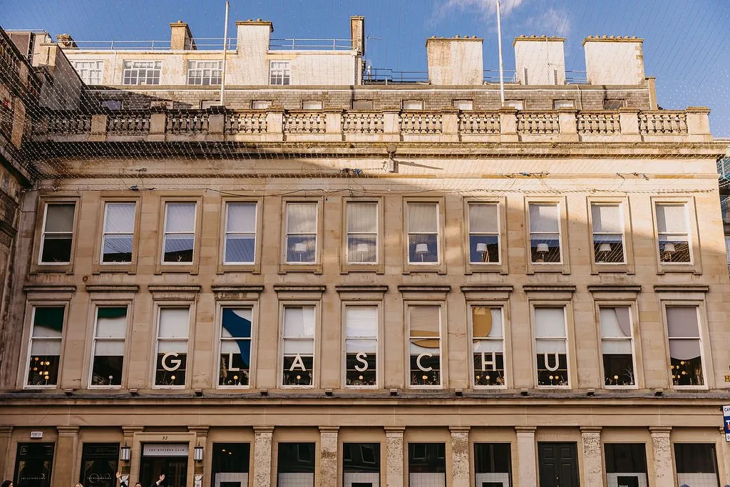This is an exterior shot of a grand, multi-story building, likely a hotel or club, constructed from light-toned stone with classical architectural details. The facade features numerous large sash windows, with the second floor prominently displaying illuminated letters spelling "GLASCHU" within the window frames, creating a striking visual. A delicate net of fairy lights is draped across the upper portion of the building, adding a festive and welcoming ambiance, even under a bright blue sky. Glimpses of interior lighting and simple lamps can be seen through some windows, hinting at the warm spaces within.