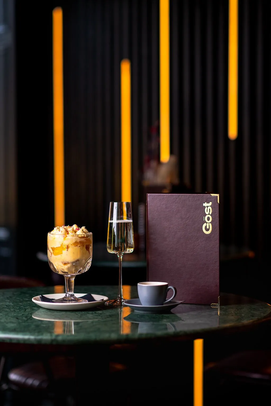 A sophisticated close-up captures a round, polished dark green marble table set for an evening experience. On it rests a decadent trifle, a flute of champagne, a minimalist grey coffee cup, and a deep burgundy menu with gold lettering, all reflecting the warm glow. The background features a dark, vertically ribbed wall accented by several striking, elongated orange light strips, creating an intimate and luxurious ambiance.