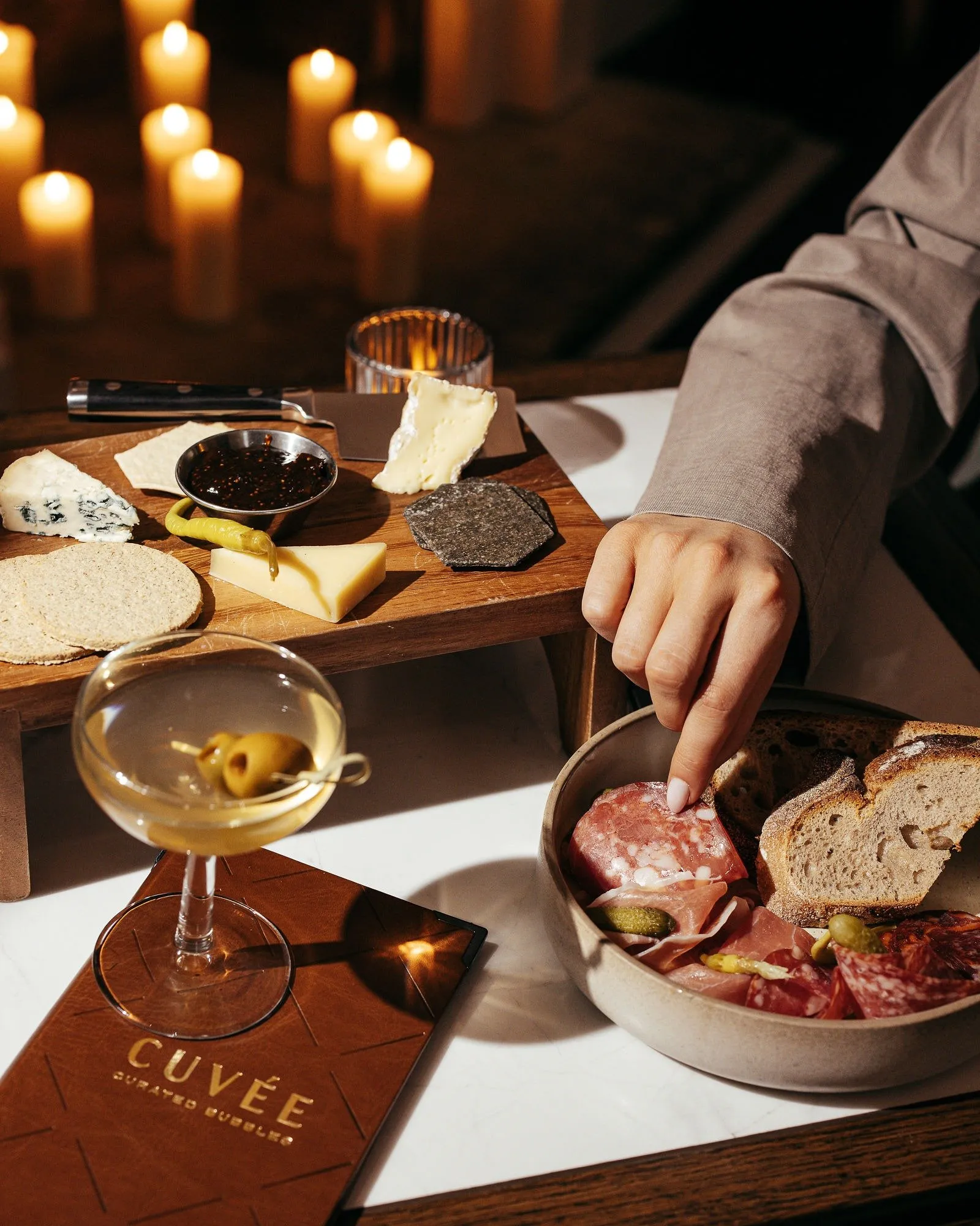 This intimate close-up captures a luxurious evening setting, centered on a white marble tabletop with dark wood accents. A curated selection of charcuterie and cheeses is artfully presented on a raised wooden board and a ceramic bowl, alongside a classic martini. The scene is bathed in the warm, soft glow of numerous flickering candles in the background, creating a cozy and sophisticated atmosphere perfect for an upscale lounge or wine bar. A person's hand reaches into the charcuterie bowl, inviting interaction with the spread, while a branded leather menu hints at a premium beverage offering.