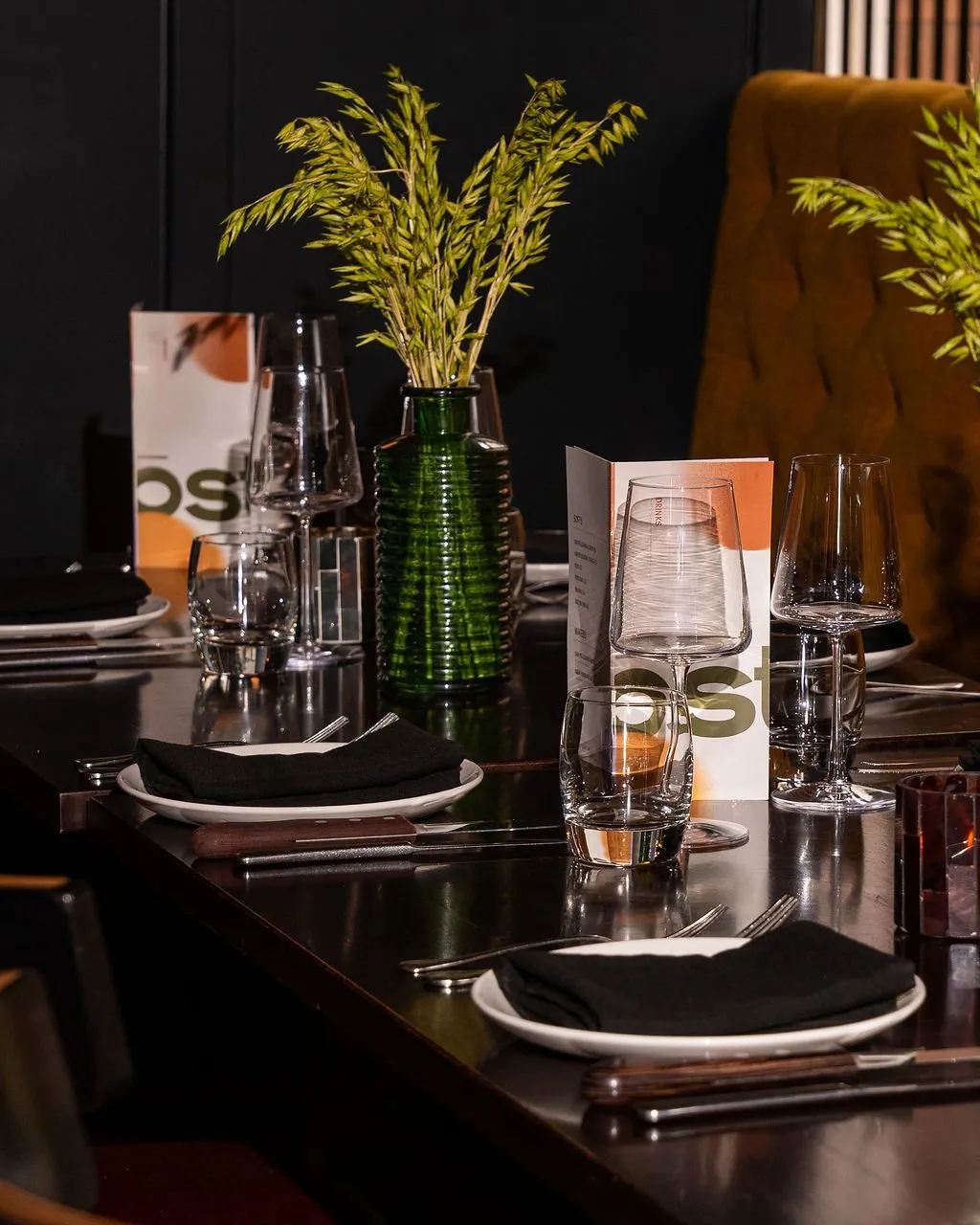 A sophisticated dining table is meticulously set for an evening meal, featuring dark polished wood, crisp white plates, and black fabric napkins. Elegant clear glassware and cutlery with dark handles complement a ribbed dark green vase holding dried golden grasses, all bathed in warm, intimate lighting that highlights the rich textures and creates a luxurious, inviting atmosphere. A glimpse of a plush, tufted mustard-gold chair and dark slatted wall paneling in the background hints at a refined restaurant interior.