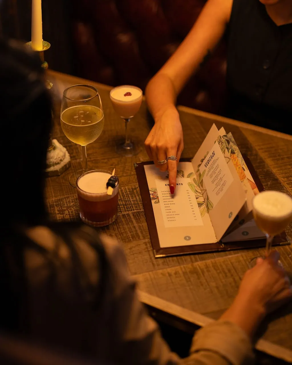 This intimate, dimly lit scene captures two patrons at a rustic wooden table, engrossed in a beautifully illustrated menu. The warm glow of a nearby candle and the rich, blurred leather upholstery in the background create a cozy, inviting atmosphere. Several artfully presented cocktails sit on the distressed timber surface, suggesting a sophisticated yet relaxed bar or lounge setting, with a focus on the tactile experience of choosing drinks.