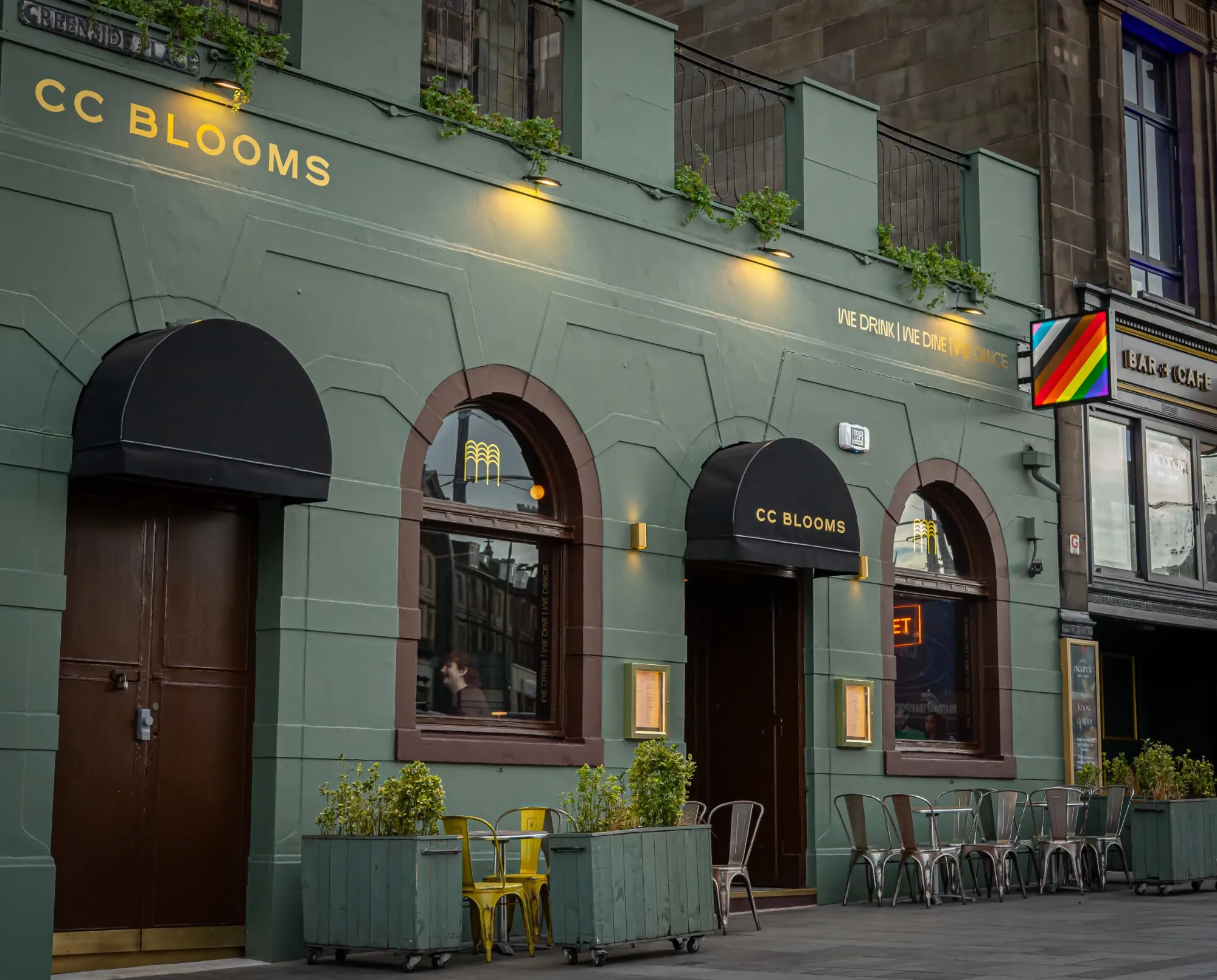 This exterior shot captures the inviting facade of "CC BLOOMS" at dusk, a hospitality venue painted in a sophisticated sage green. Gold lettering and warm spotlights illuminate the arched windows and entrances, which are topped with sleek black dome awnings. A casual outdoor seating area features vibrant yellow and distressed metal chairs alongside mobile wooden planters, creating a welcoming street-level presence.