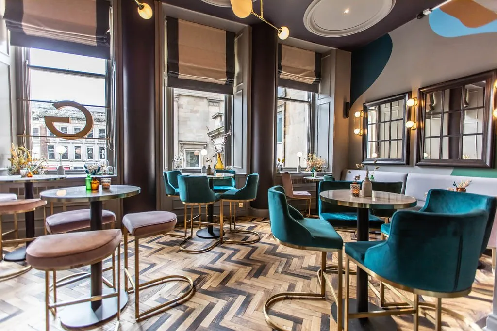 This vibrant hospitality space features a mix of high-top and banquette seating, bathed in natural light from tall windows and complemented by warm, sculptural artificial lighting. The design blends rich teal velvet and blush upholstery with brass accents, set against a striking herringbone wood floor and walls adorned with an abstract, multi-toned mural. The layout encourages both intimate conversations and lively socialising, creating a sophisticated yet playful atmosphere.