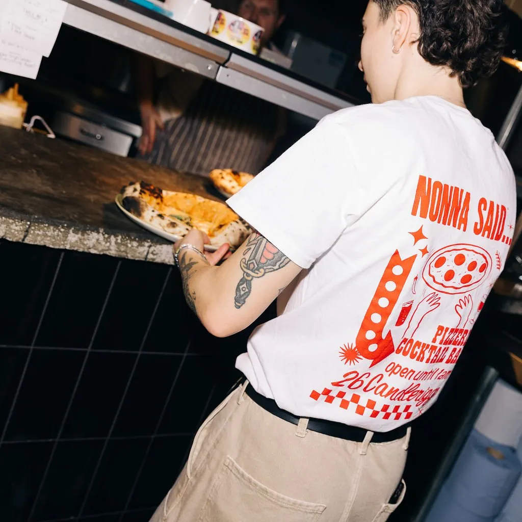 This image captures a lively kitchen pass-through area, focusing on a server placing a freshly baked, charred-crust pizza onto a rugged, dark stone counter. The backdrop features dark, square-tiled walls and a stainless steel shelf, with a chef partially visible in the warm, focused lighting that highlights the food and the bustling, authentic atmosphere. The server's casual uniform, a white t-shirt with bold red graphics, adds a touch of modern, playful branding to the functional space.