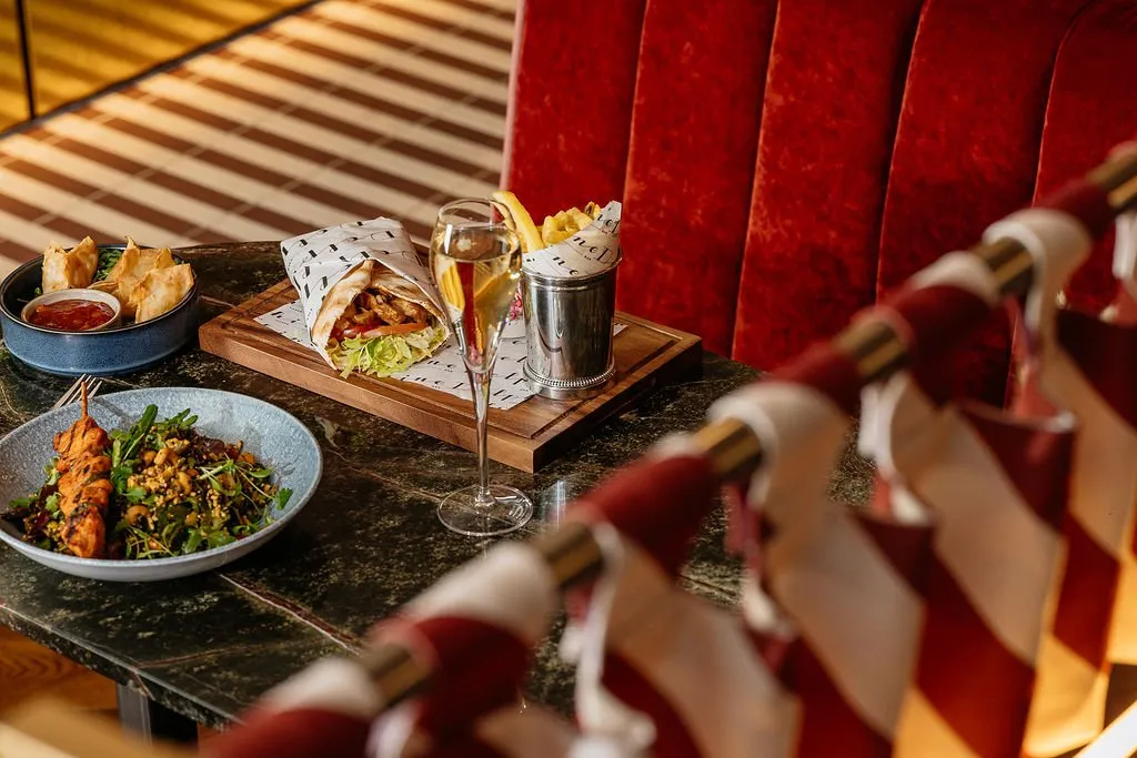 A close-up shot of a dark marble table set with various dishes and a champagne flute, against a backdrop of a luxurious red velvet banquette and a striped floor.