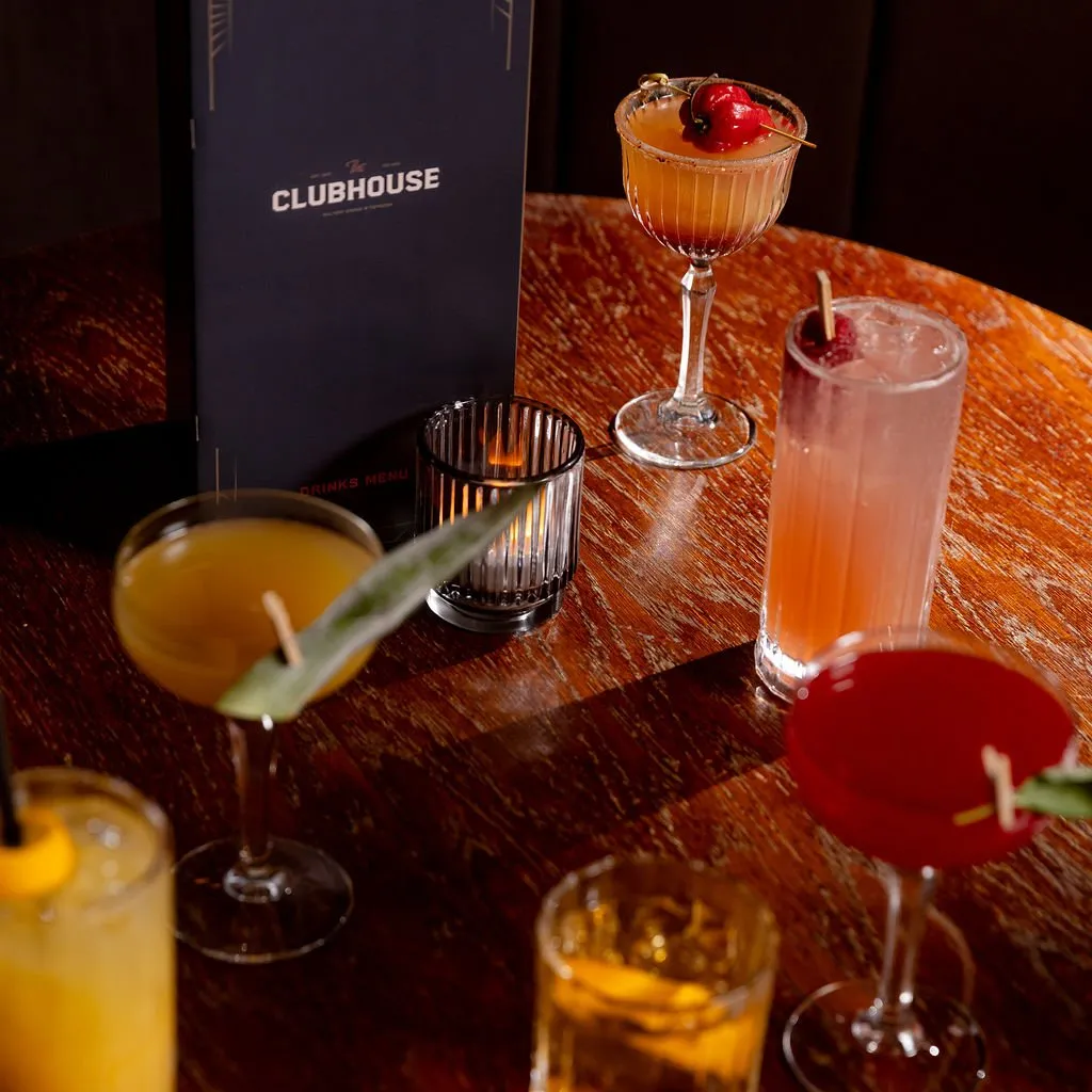 This intimate shot captures a rich, dark wooden table laden with an array of vibrant cocktails, a sleek "The Clubhouse" menu, and a flickering candle in a fluted glass holder. The warm, low lighting creates a sophisticated and inviting atmosphere, highlighting the drinks' colors and the table's prominent wood grain. It suggests a cozy, upscale lounge or bar setting perfect for evening gatherings. The composition draws attention to the curated details and the overall luxurious mood.