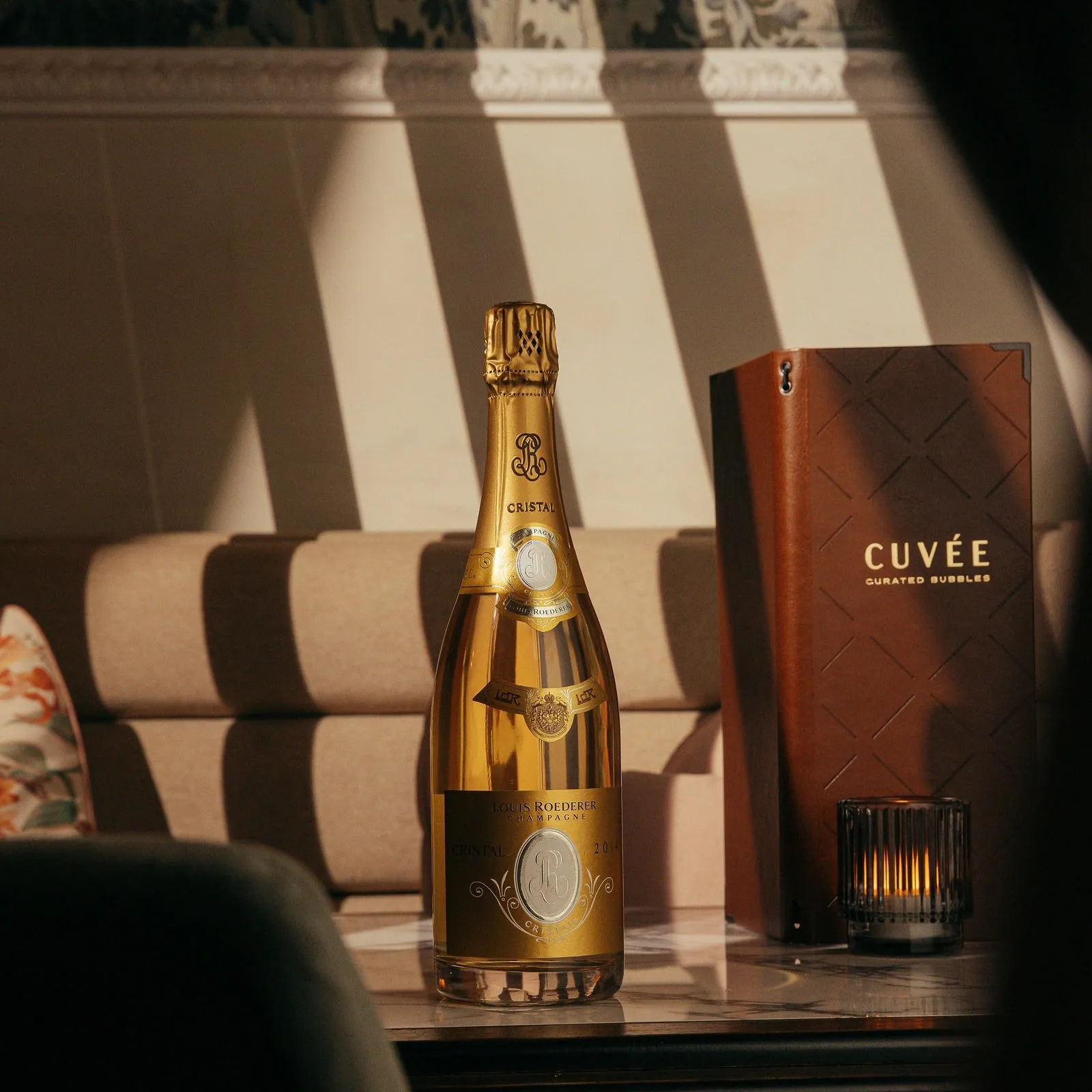 This close-up shot captures a luxurious table setting, featuring a gleaming bottle of Louis Roederer Cristal champagne, a rich brown leather "Cuvée" menu with a subtle diamond pattern, and a fluted glass votive candle holder casting a warm glow. Dramatic natural light creates strong diagonal shadows across the beige upholstered banquette seating and a classic decorative wall molding in the background. The scene evokes an intimate and sophisticated mood, highlighting opulent details and refined finishes.
