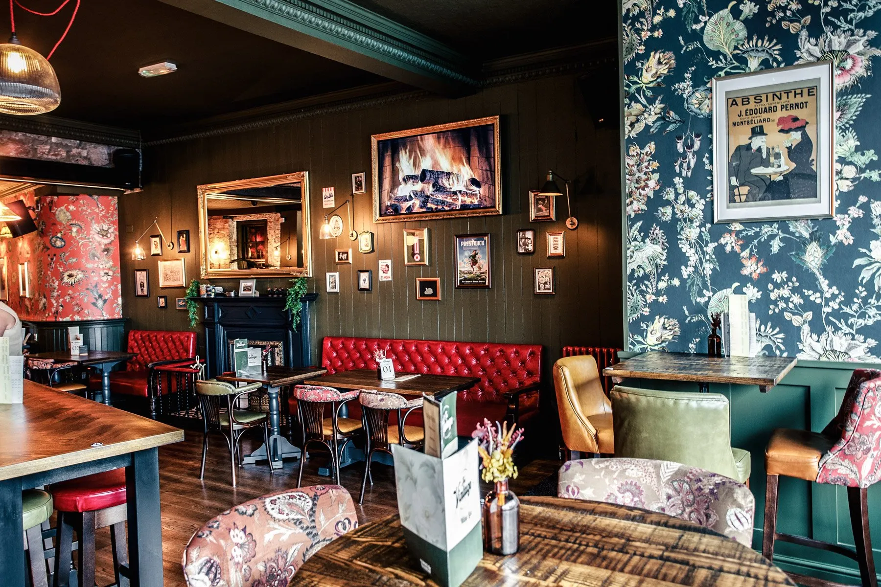 This inviting hospitality space features a rich, eclectic design with deep jewel tones and varied textures. Dark green paneled walls are adorned with a dense gallery wall, including a prominent digital fireplace and a large ornate mirror, while sections are enlivened by vibrant floral wallpapers in red and dark blue. Plush red tufted banquettes offer comfortable seating alongside an assortment of wooden and upholstered chairs, all bathed in warm, ambient lighting that creates an intimate and traditional pub-like atmosphere.