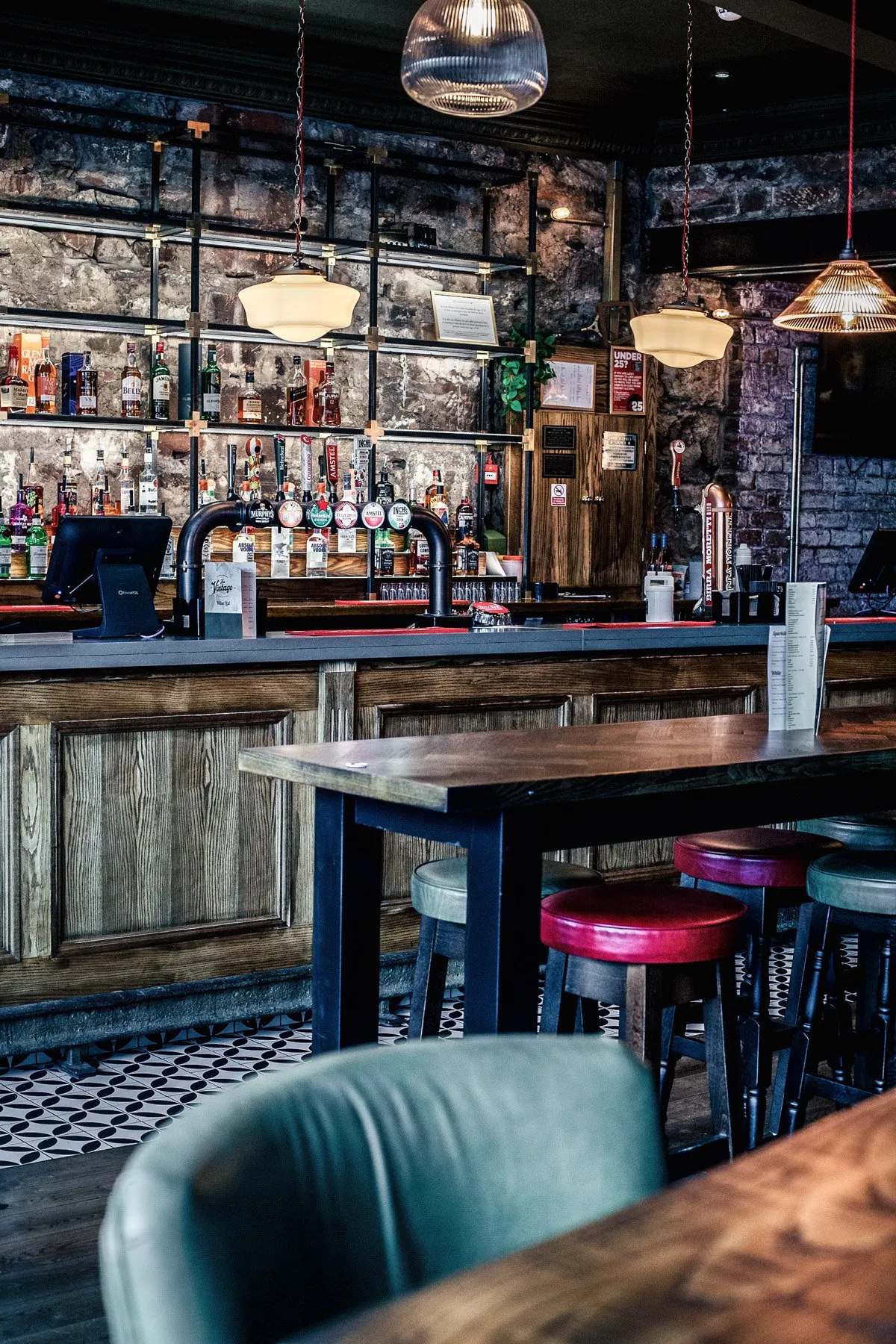 This inviting bar interior features a rich blend of rustic and industrial elements, with exposed dark brick walls and a dark wood-paneled bar front creating a cozy, intimate atmosphere. A striking metal shelving unit behind the bar, filled with liquor bottles, is illuminated by warm backlighting and various pendant lights, including fluted glass and opaque globes. High wooden tables with dark metal bases are paired with an eclectic mix of bar stools, some upholstered in vibrant red, purple, and green leather, complementing the geometric black and white patterned floor tiles.