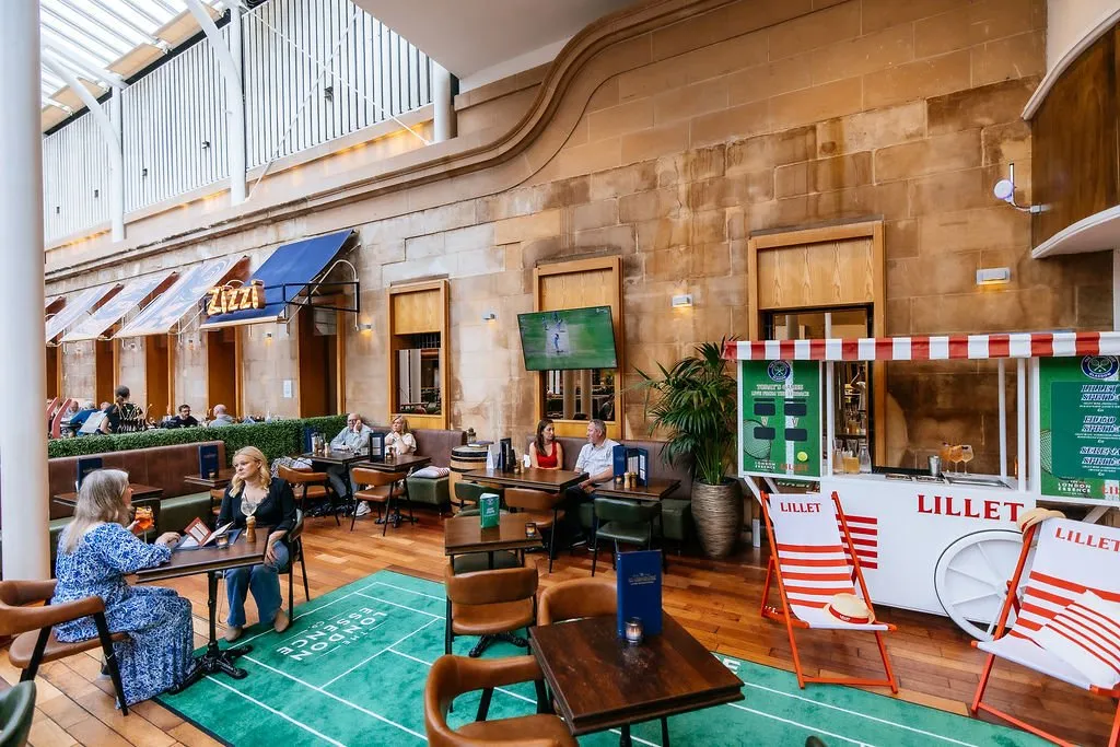This image captures a vibrant, high-ceilinged indoor hospitality space, likely an atrium or covered arcade, featuring a long, impressive wall of exposed warm-toned stone masonry. The area is laid out with a mix of dark leather banquette seating and freestanding dark wood tables with upholstered chairs, all bathed in bright natural light from above, complemented by subtle wall sconces. A playful, temporary Lillet pop-up bar with red and white striped awnings, tennis-themed branding, and matching deck chairs adds a lively, event-driven atmosphere, contrasting with the grand architectural backdrop.