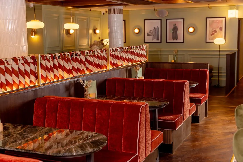 A vibrant and inviting restaurant interior features rows of red velvet banquettes and dark marble tables, set against sage green paneled walls.