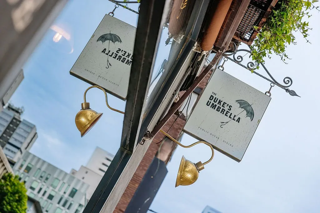 The image captures a low-angle view of "The Duke's Umbrella" public house and dining signage, featuring two identical, distressed white signs with a black umbrella logo and text. Each sign is illuminated by a gooseneck brass wall light, mounted on an ornate black wrought-iron bracket. A vibrant green vine trails from an upper window box, adding a touch of nature, while the reflection in a large window reveals the reverse side of the sign and the modern urban skyline beyond, creating a dynamic contrast between classic pub aesthetics and contemporary city architecture.