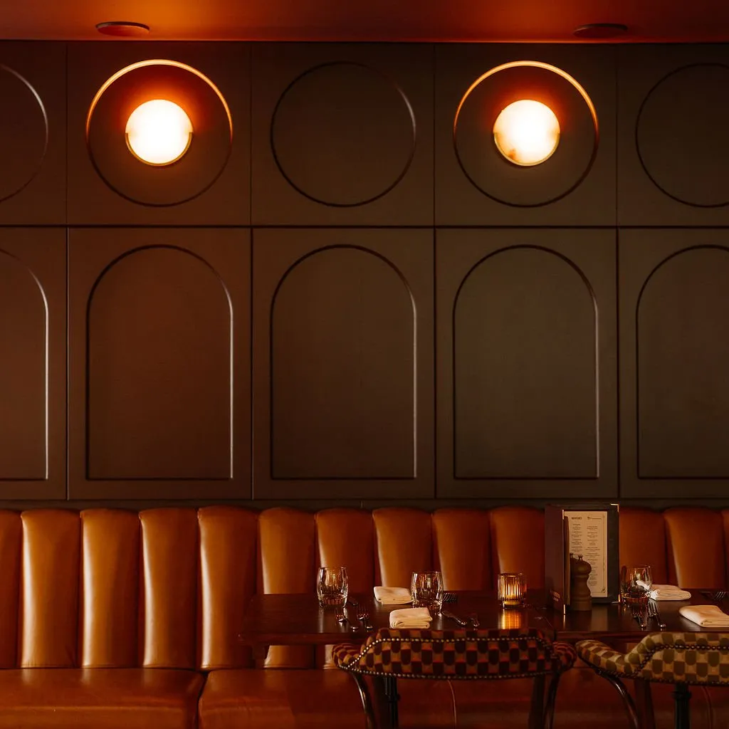This intimate dining space features a sophisticated palette of deep, muted tones and warm leather. A dark paneled wall, adorned with a grid of recessed arches and circular cutouts housing glowing sconces, creates a dramatic backdrop. A long, vertically tufted cognac leather banquette provides plush seating, complemented by dark wood tables set for dinner and chairs upholstered in a subtle checkered pattern. The ambient, warm lighting casts a cozy, inviting glow, enhancing the classic yet modern atmosphere.
