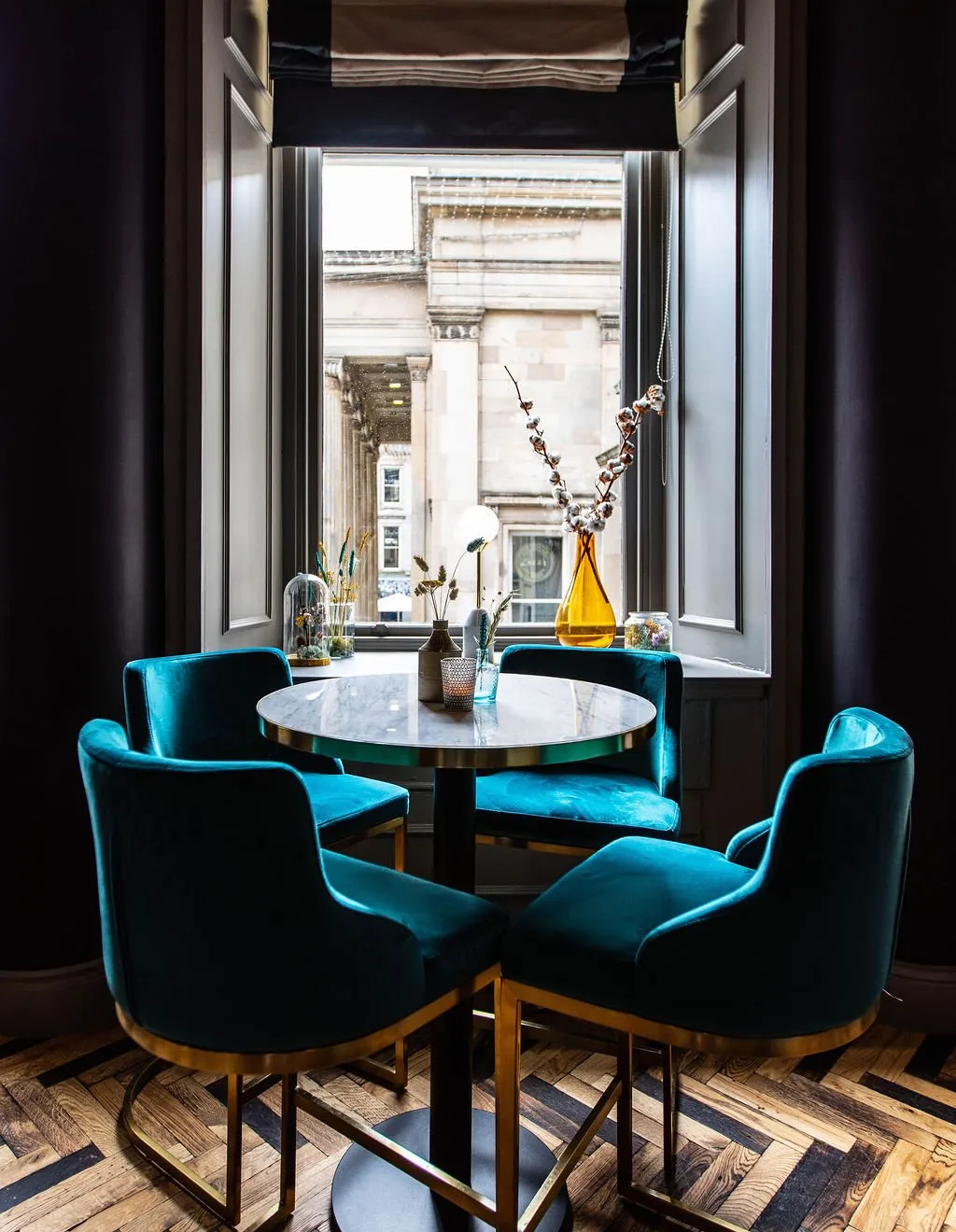 This intimate window nook features a round marble-topped table with a brass edge, surrounded by four plush teal velvet bar stools with elegant brass frames and integrated footrests, set against a dark, moody backdrop. Natural light streams in from the large window, illuminating decorative dried botanicals and a modern table lamp on the sill, while a herringbone wood floor adds warmth and texture to the sophisticated, slightly dramatic ambiance. The view outside reveals a grand classical building, grounding the space in its urban context.