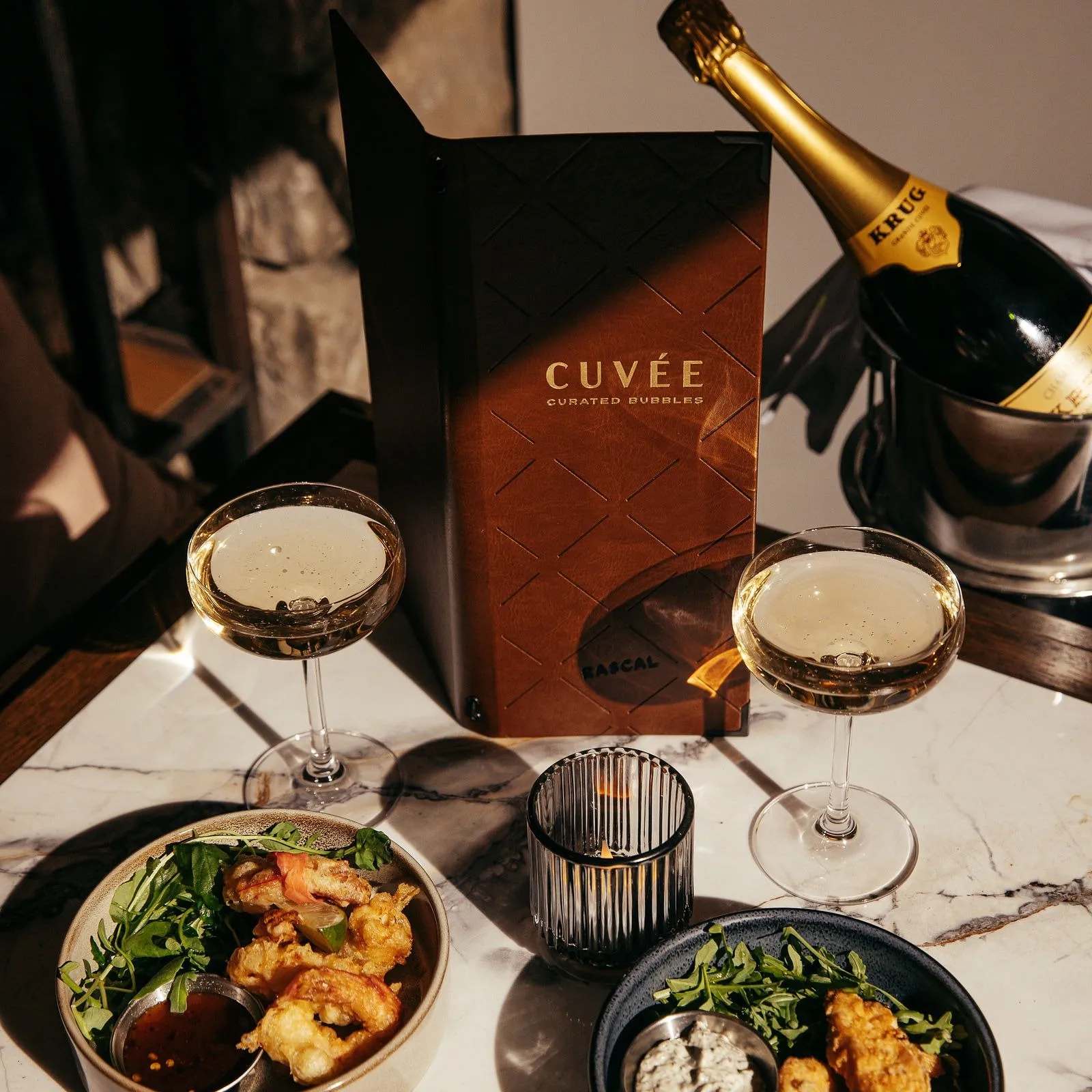This intimate close-up captures a luxurious table setting for two, featuring two coupe glasses filled with sparkling wine, a bottle of Krug champagne in a sleek ice bucket, and two elegant bowls of appetizers. The scene is set on a veined white marble tabletop, illuminated by warm, directional light that casts soft shadows and highlights the rich textures of the brown leather menu and the fluted glass votive candle holder, creating a sophisticated and inviting atmosphere. In the background, hints of a rough stone wall suggest a refined, perhaps historic, interior.