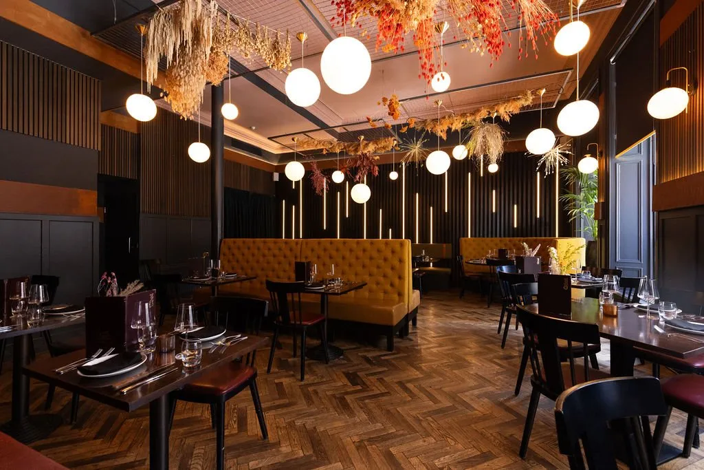 This sophisticated restaurant interior features a warm, inviting ambiance created by a rich material palette and layered lighting. Dark wood slatted walls and classic paneling provide a dramatic backdrop for plush, mustard-yellow tufted banquettes and dark wooden tables with mixed seating. Overhead, a striking installation of dried botanicals in warm autumnal hues is interspersed with numerous spherical pendant lights, casting a soft, intimate glow across the herringbone parquet floor, complemented by vertical LED strips on the walls.