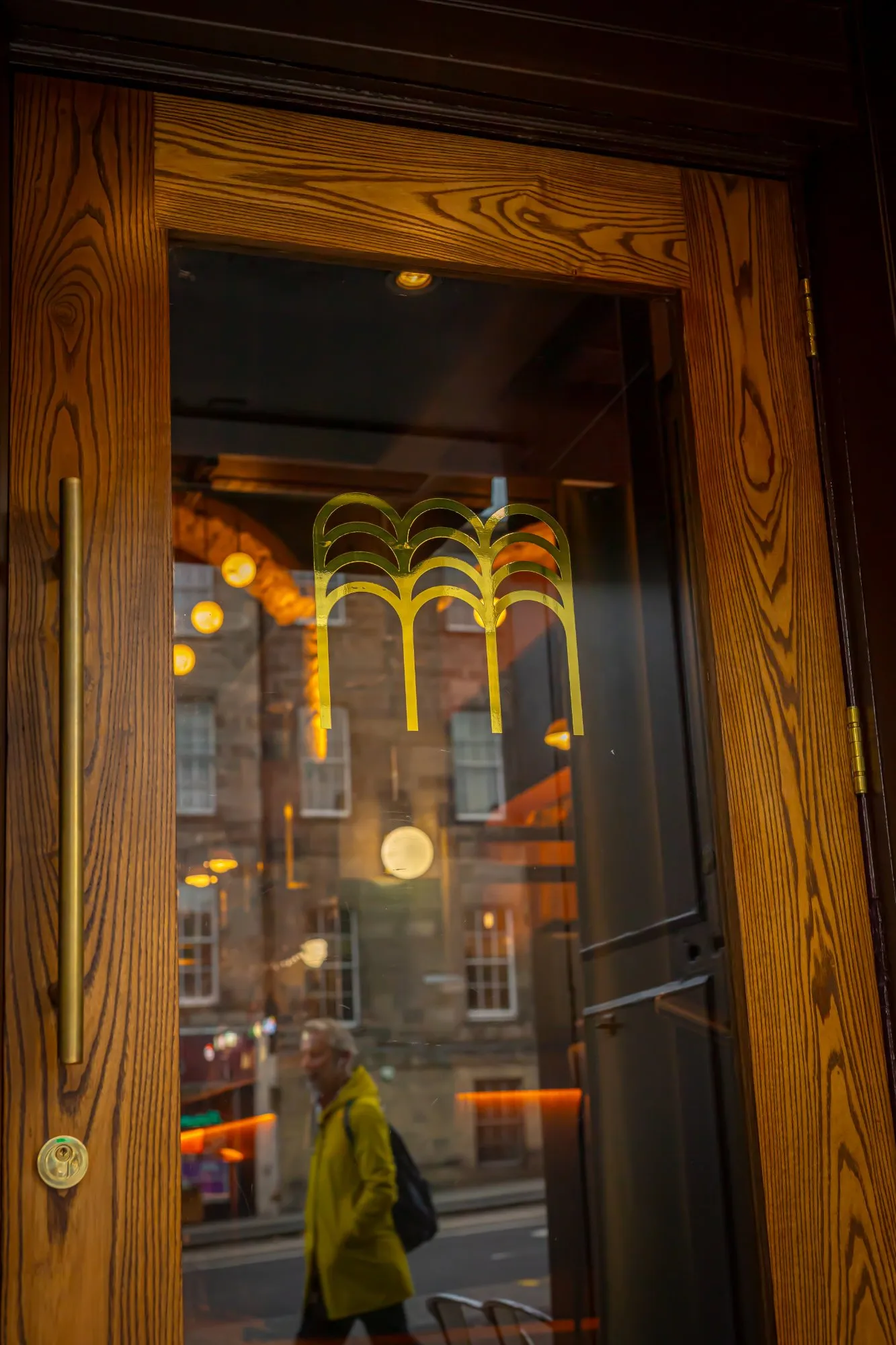 This image captures the sophisticated entrance to a hospitality venue, showcasing a rich, warm-toned wooden door with a prominent, natural grain. A large glass panel is elegantly adorned with a distinctive gold, layered arch motif logo, which subtly reflects both the inviting warm glow of the interior lighting and the blurred, dynamic urban streetscape beyond. The door is completed with a sleek, elongated brass pull handle and a matching brass lock, creating a refined and welcoming first impression.