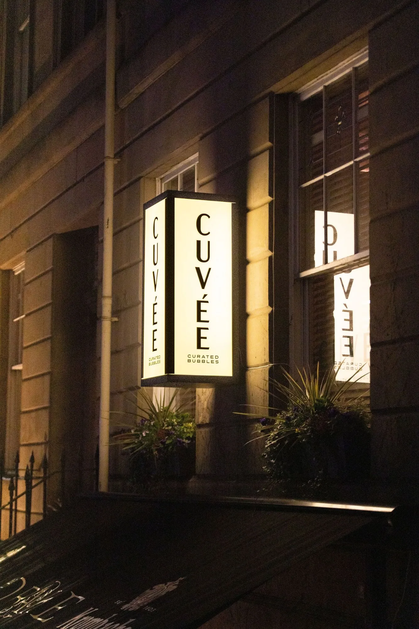 This exterior shot captures the inviting facade of 'CUVÉE Curated Bubbles' at night, highlighted by a prominent, vertically oriented illuminated sign with a warm, creamy glow. The building features a dark, classic stone or stucco facade and traditional multi-pane windows, with lush planter boxes adding a touch of natural texture below. The warm light from the signage and subtle interior reflections creates a sophisticated and welcoming nocturnal atmosphere, hinting at an upscale beverage experience within.