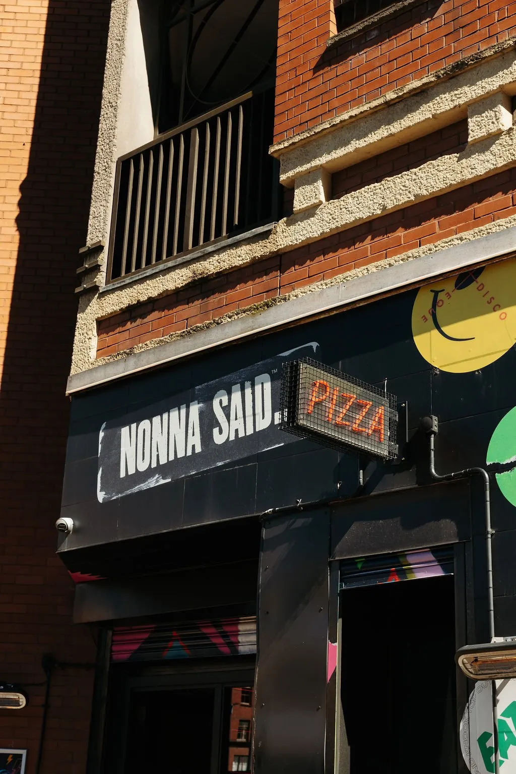 The image captures the vibrant, multi-layered facade of an urban hospitality venue, "NONNA SAID. PIZZA." The upper floors feature traditional red brickwork with textured, light-colored rendered bands and a dark metal balcony railing, casting sharp shadows under direct sunlight. Below, the ground floor is clad in sleek black panels, providing a stark contrast and a modern backdrop for the prominent white "NONNA SAID." signage and a glowing orange LED "PIZZA" sign. Playful yellow and green disco-themed smiley face graphics adorn the black facade, hinting at a lively atmosphere, while the partially open roller shutters reveal abstract pink and blue geometric patterns, adding a touch of edgy, retro flair to the entrance.