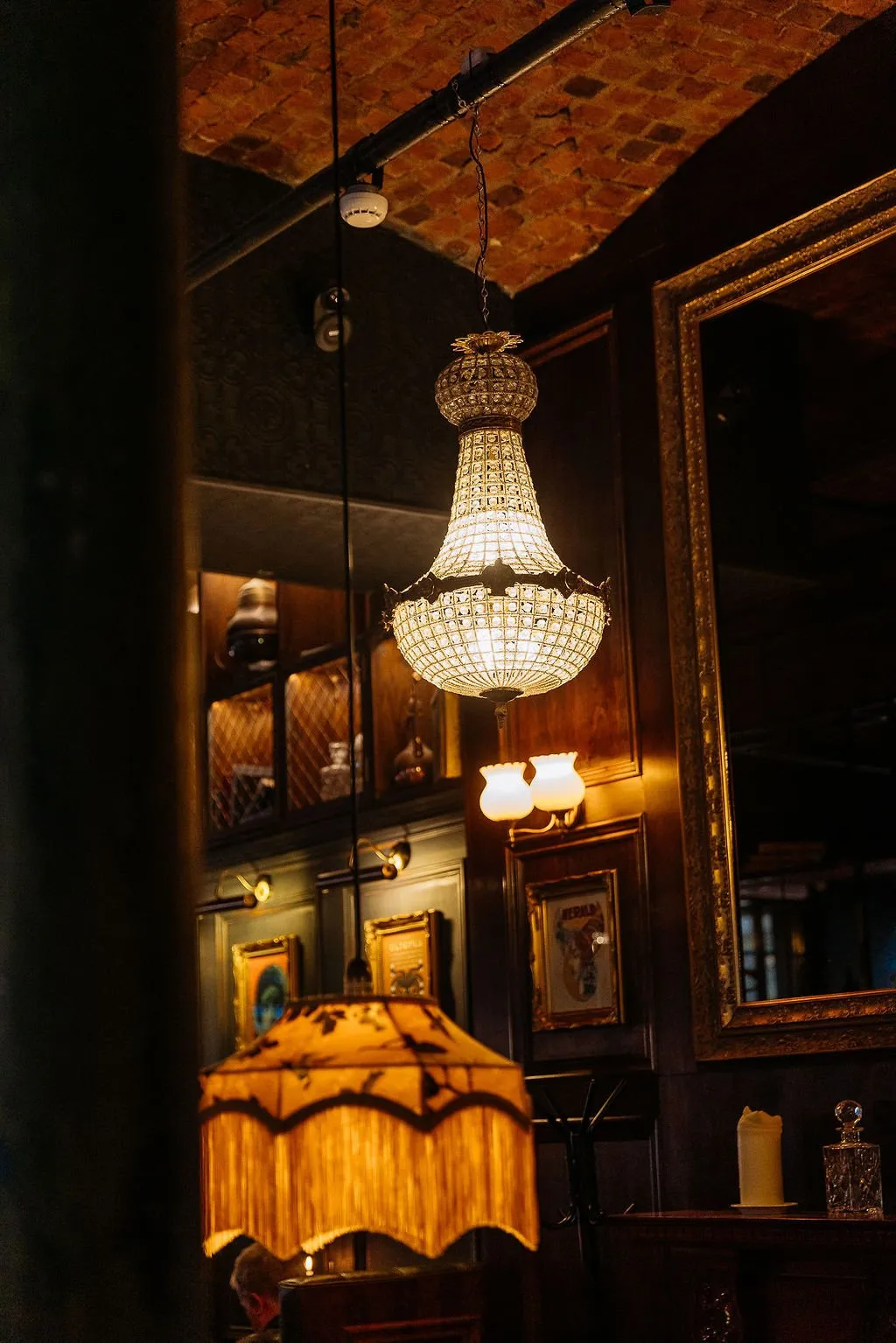 This intimate interior features a rich, layered design with warm, ambient lighting. A grand crystal chandelier hangs prominently from an exposed brick ceiling, contrasting with dark wood paneling and ornate framed artwork on the walls. A fringed table lamp casts a soft glow, contributing to the cozy, opulent, and slightly moody atmosphere, perfect for a sophisticated lounge or bar setting. The space blends traditional elegance with a hint of industrial texture from the exposed brick.