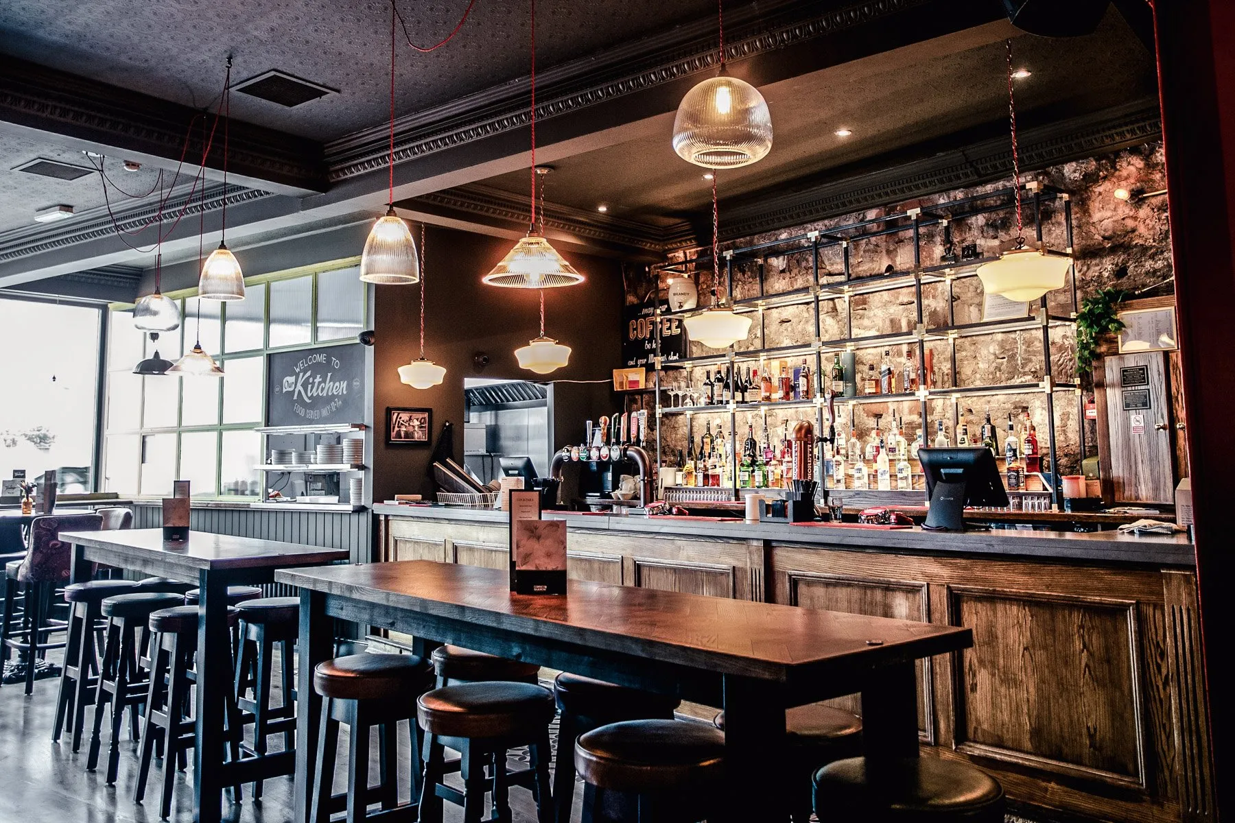 This hospitality space features a moody, industrial-rustic aesthetic, anchored by a long, dark wooden bar with paneling and a striking exposed stone wall backdrop. Overhead, a mix of ribbed glass and opaque bowl-shaped pendant lights with vibrant red fabric cords cast a warm, inviting glow, complementing the natural light filtering through large, textured glass windows. High communal tables with dark leather-topped stools provide casual seating, creating an intimate yet functional environment perfect for dining and socializing.