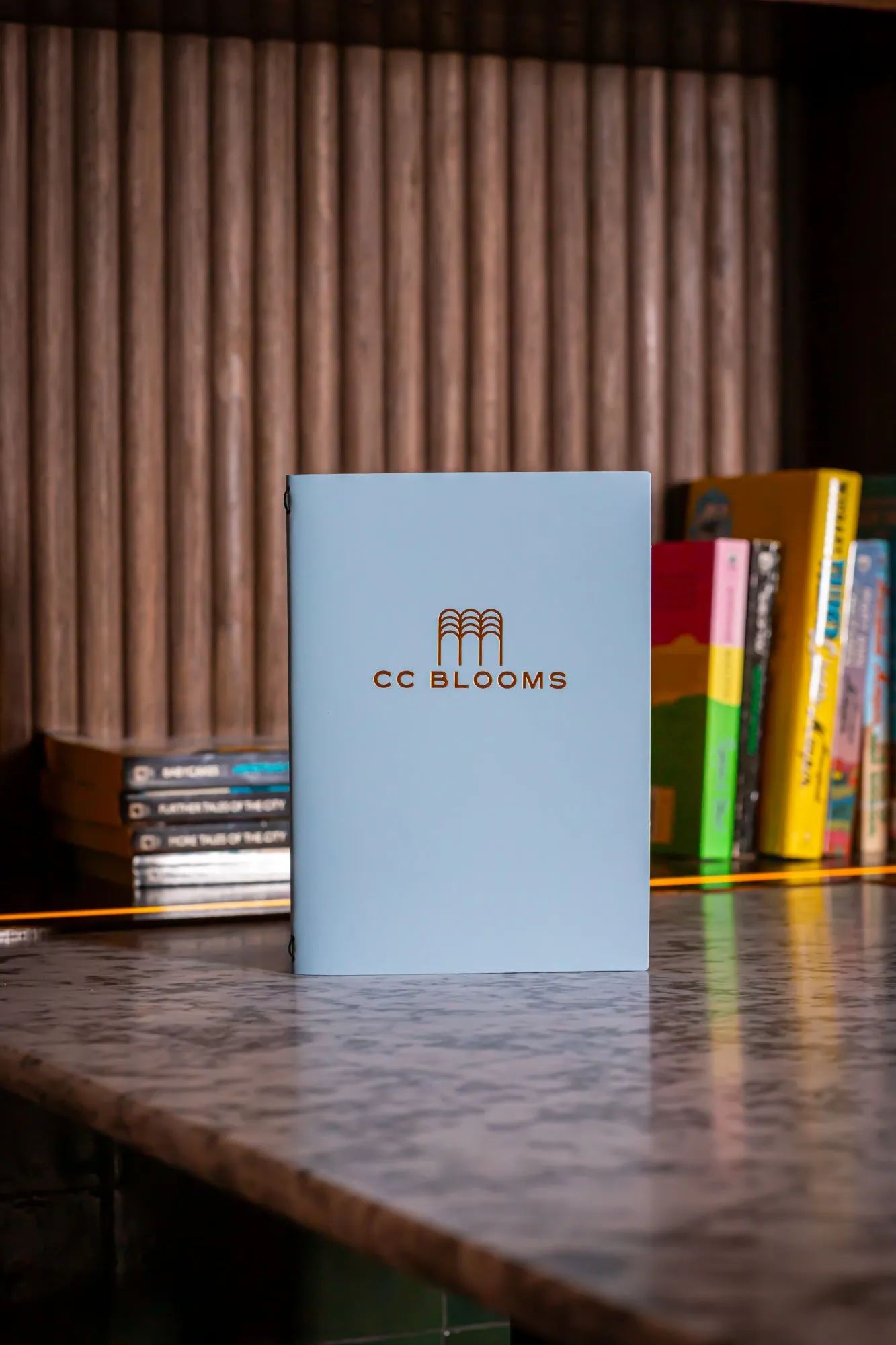 This detail shot captures a light blue menu with a copper 'CC BLOOMS' logo, standing prominently on a polished, veined marble counter. In the background, a warm, fluted wooden wall or shelving unit is subtly lit by an orange LED strip, showcasing a curated collection of books. The overall mood is sophisticated and inviting, highlighting tactile materials and thoughtful branding within a cozy, intimate setting.