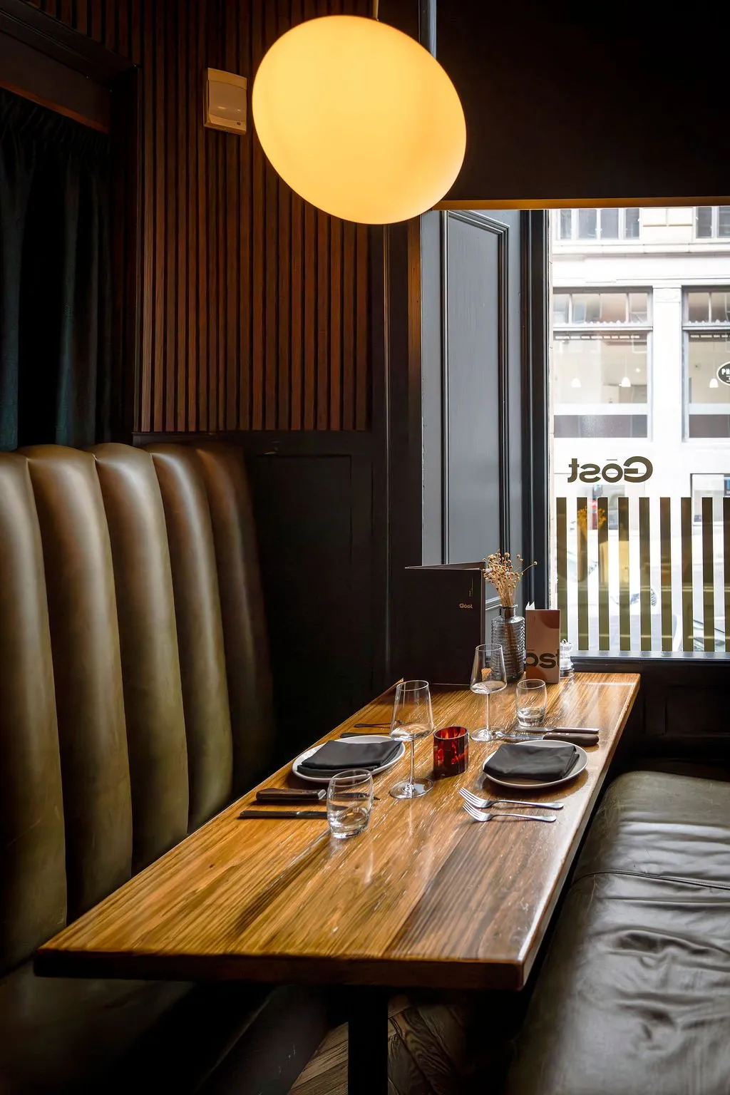 This inviting restaurant booth features a long, polished dark wood table flanked by two banquettes: a high-backed, channel-tufted olive leather seat against a dark wood slatted wall, and a simpler, lower leather bench. A large, spherical opaque pendant light casts a warm, ambient glow, complementing the natural light filtering through the striped window overlooking a city street. The space exudes a sophisticated yet cozy mood, with thoughtful details like the elegant table settings and a small vase of dried botanicals, creating an intimate dining experience.